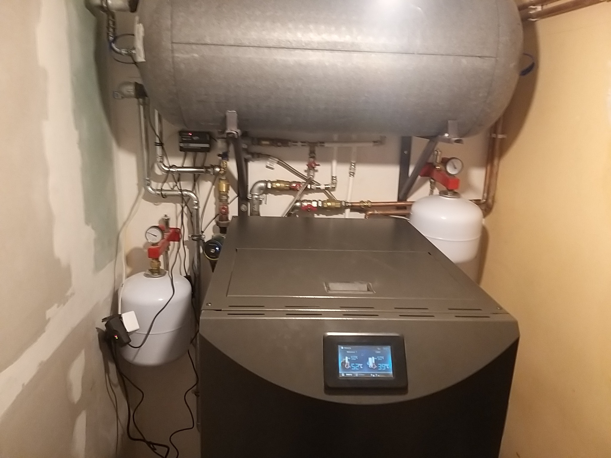 Nowoczesny piec centralnego ogrzewania z ekranem dotykowym, zbiornik akumulacyjny powyżej, instalacja rurowa z manometrami i zbiornikami wyrównawczymi w kotłowni o jasnych ścianach.