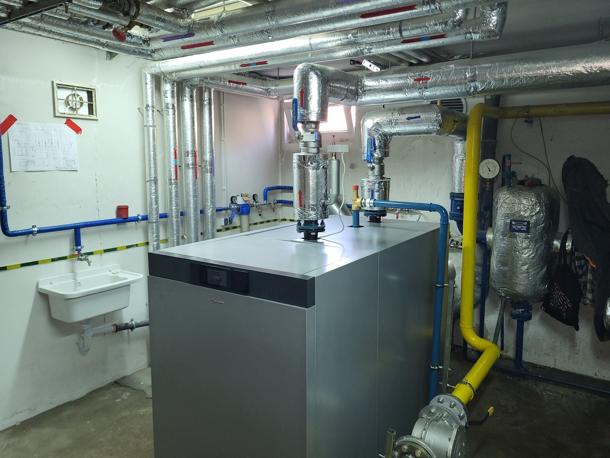 Szary piec grzewczy Viessmann w piwnicy, z widocznymi rurami instalacji grzewczej, umywalką i schematem na ścianie.