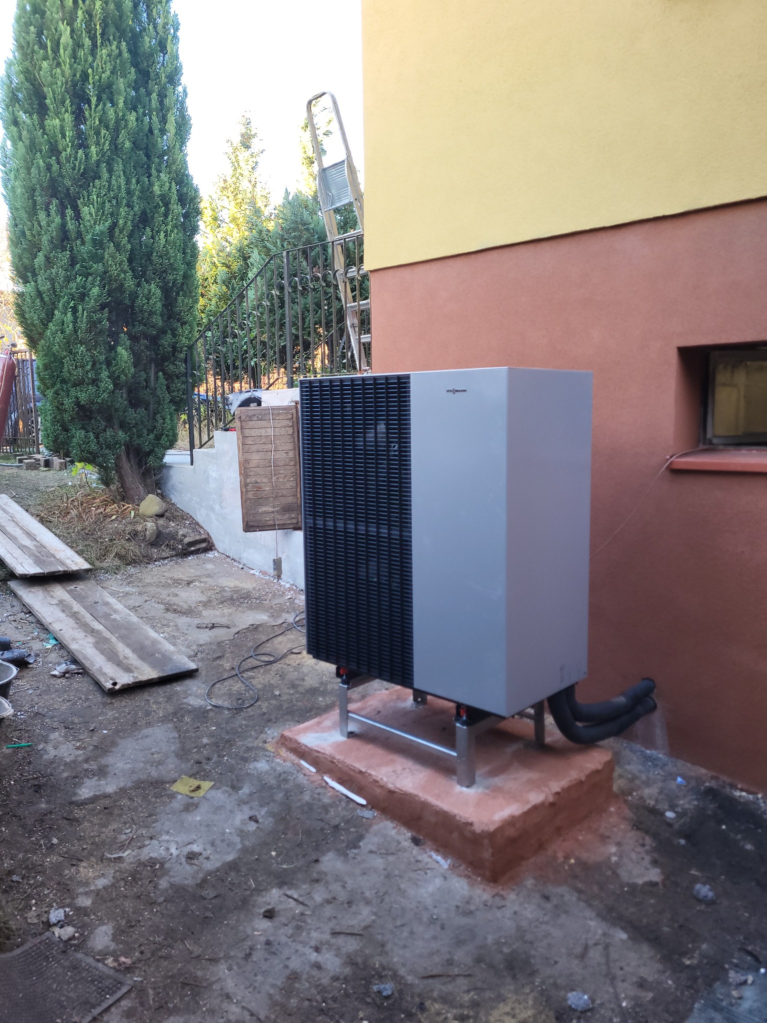 Zewnętrzna pompa ciepła Viessmann zamontowana na betonowej podstawie przy ścianie budynku, widoczne rury przyłączeniowe, drabina oparta o ogrodzenie w tle.