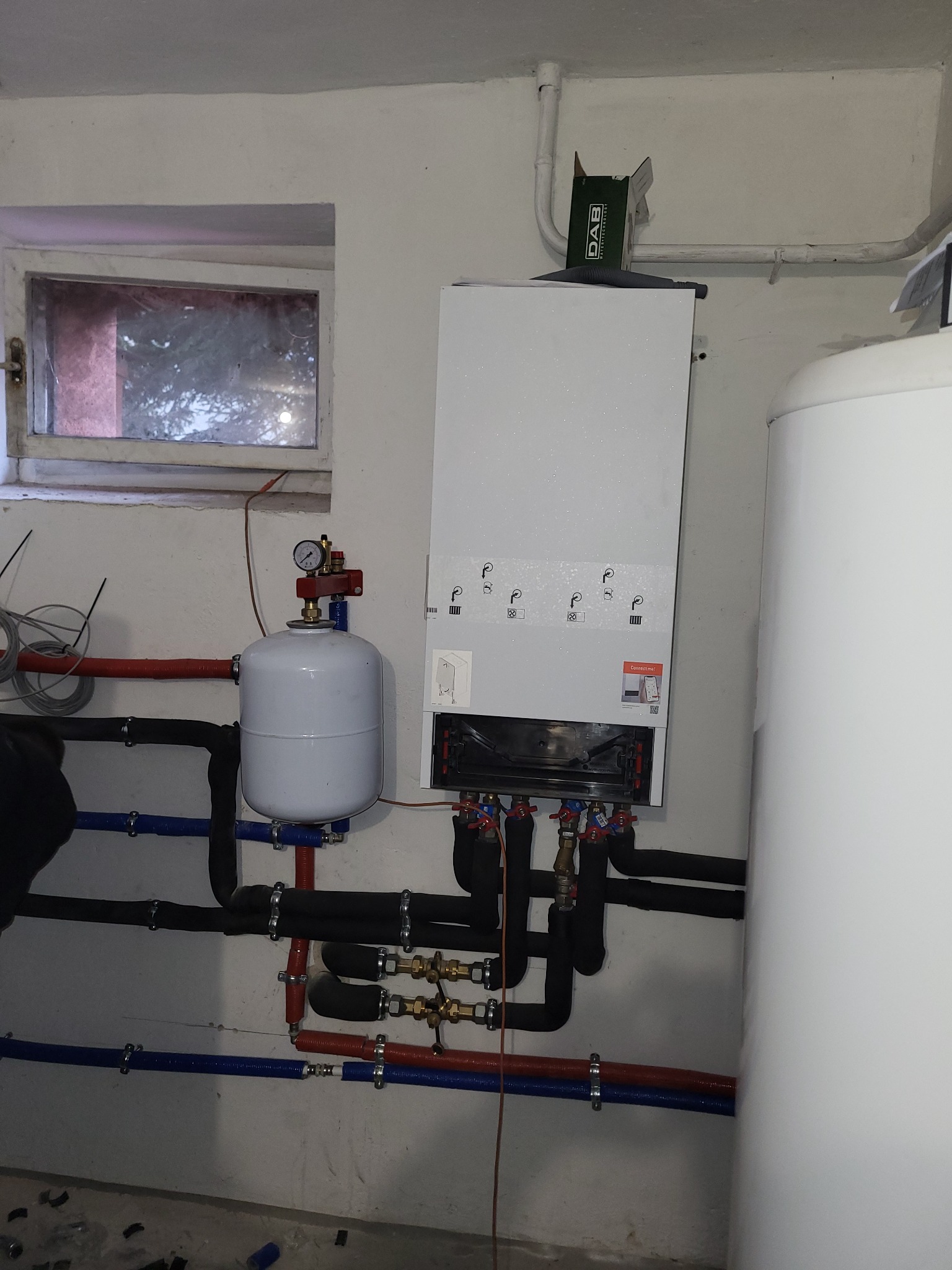Nowoczesny piec grzewczy wiszący z podłączonymi rurami w kolorach czerwonym i niebieskim oraz białym zbiornikiem wyrównawczym, zamontowany na ścianie w piwnicy w Olsztynie.