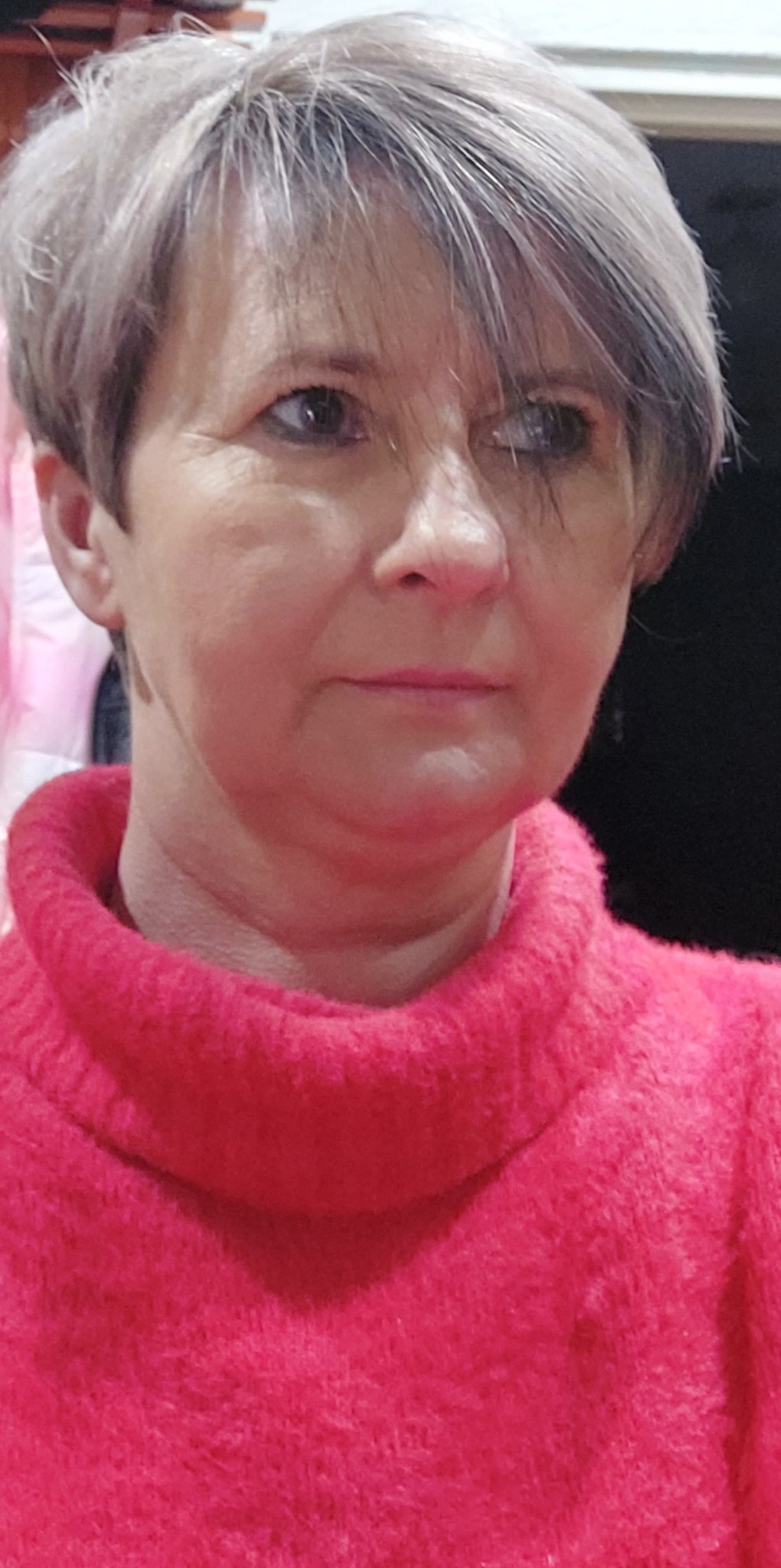 Portret kobiety z krótkimi, siwymi włosami i w różowym swetrze z golfem, patrzącej w bok.