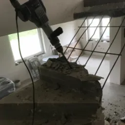 Młot udarowy Bosch w trakcie pracy nad rozbiórką betonowych schodów z wystającymi prętami zbrojeniowymi; widoczne gruz i kurz, w tle okno i elementy wykończeniowe wnętrza.