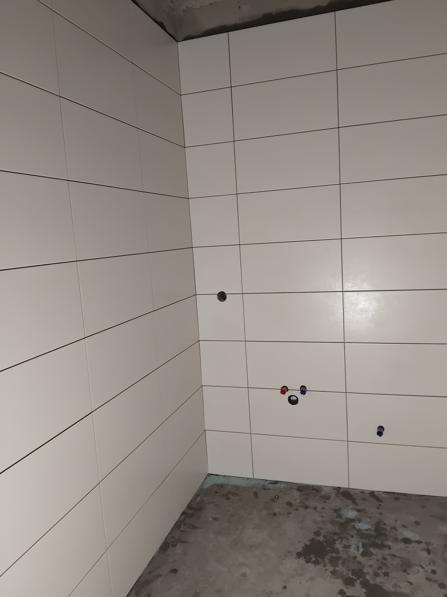 Ściany wyłożone jasnymi, kwadratowymi płytkami w łazience. Widoczne otwory na armaturę i fragment surowej podłogi. Minimalistyczny design i świeżo położone płytki.