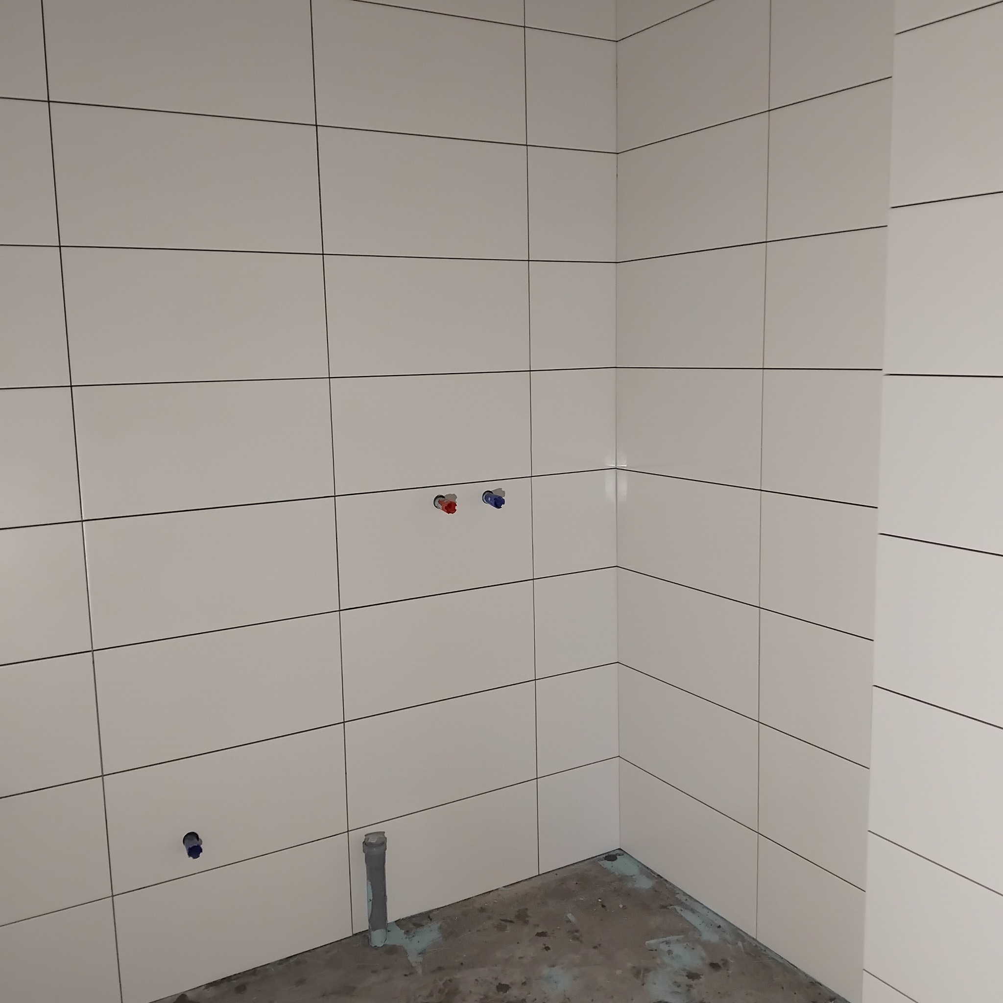 Ściany wyłożone białymi, prostokątnymi płytkami w łazience. Widoczne otwory na armaturę i rurę odpływową. Minimalistyczny design i precyzyjne ułożenie płytek.