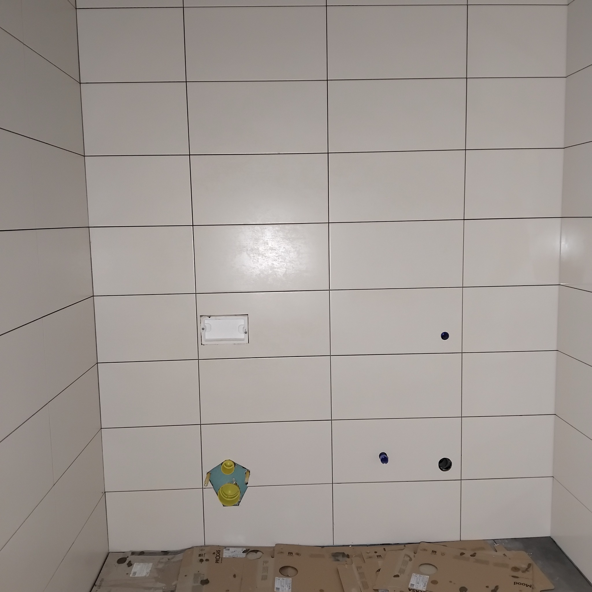 Ściana w łazience z białymi, kwadratowymi płytkami. Widoczne otwory na rury i gniazdko elektryczne. Na podłodze kartony zabezpieczające.