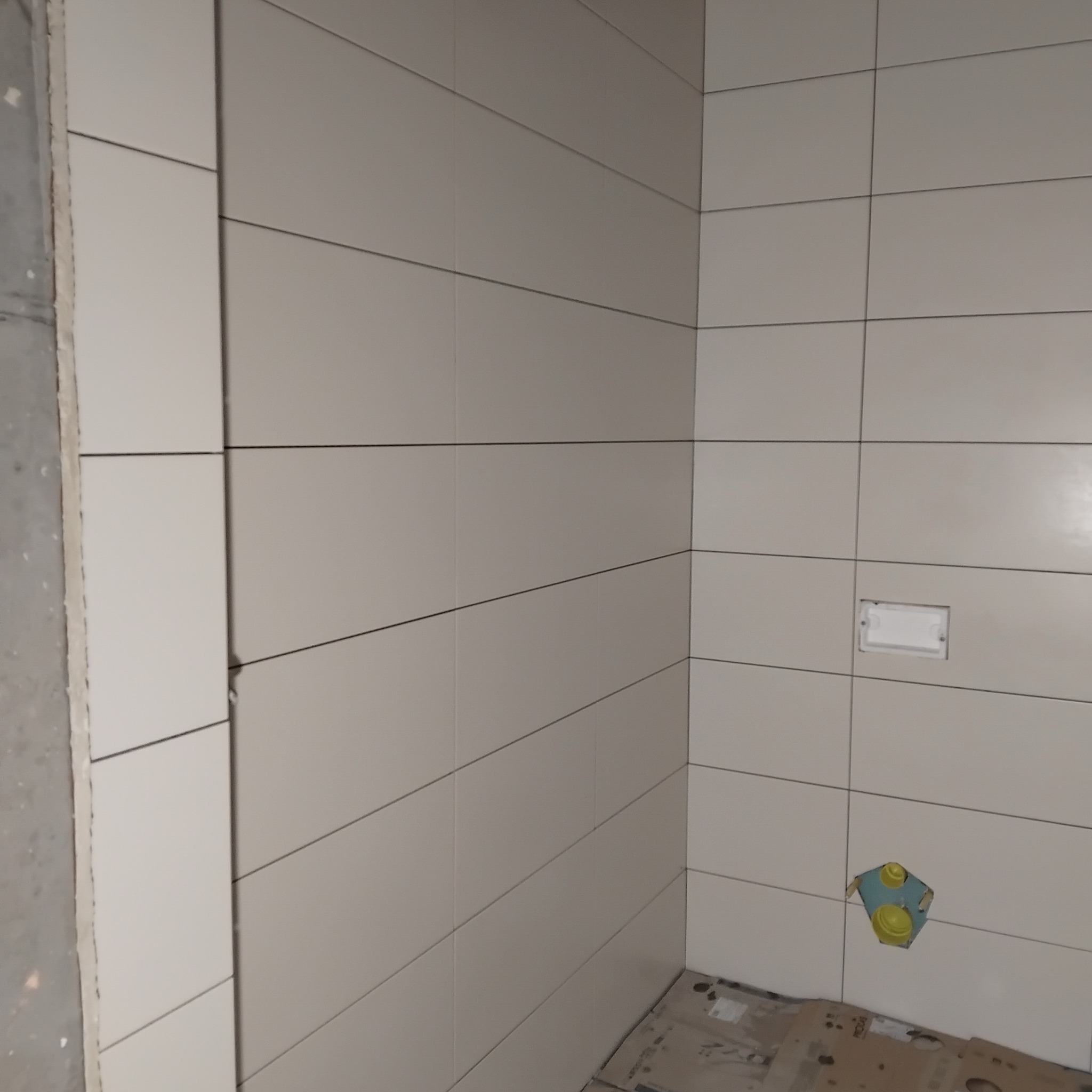 Wykończona ściana łazienki z jasnymi, prostokątnymi płytkami. Widoczny narożnik i przygotowane miejsce na armaturę. Na podłodze kartony zabezpieczające.