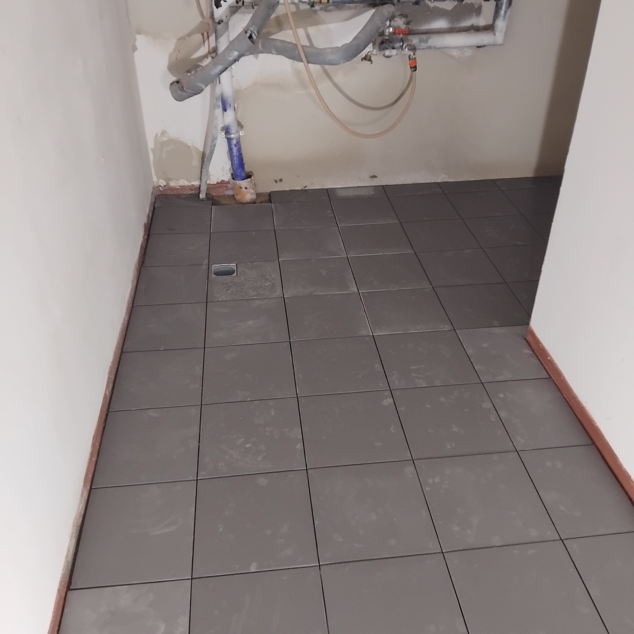 Ułożone szare płytki podłogowe w łazience z odpływem liniowym. Widoczne rury instalacji nad posadzką. Ściany przygotowane do dalszych prac wykończeniowych.