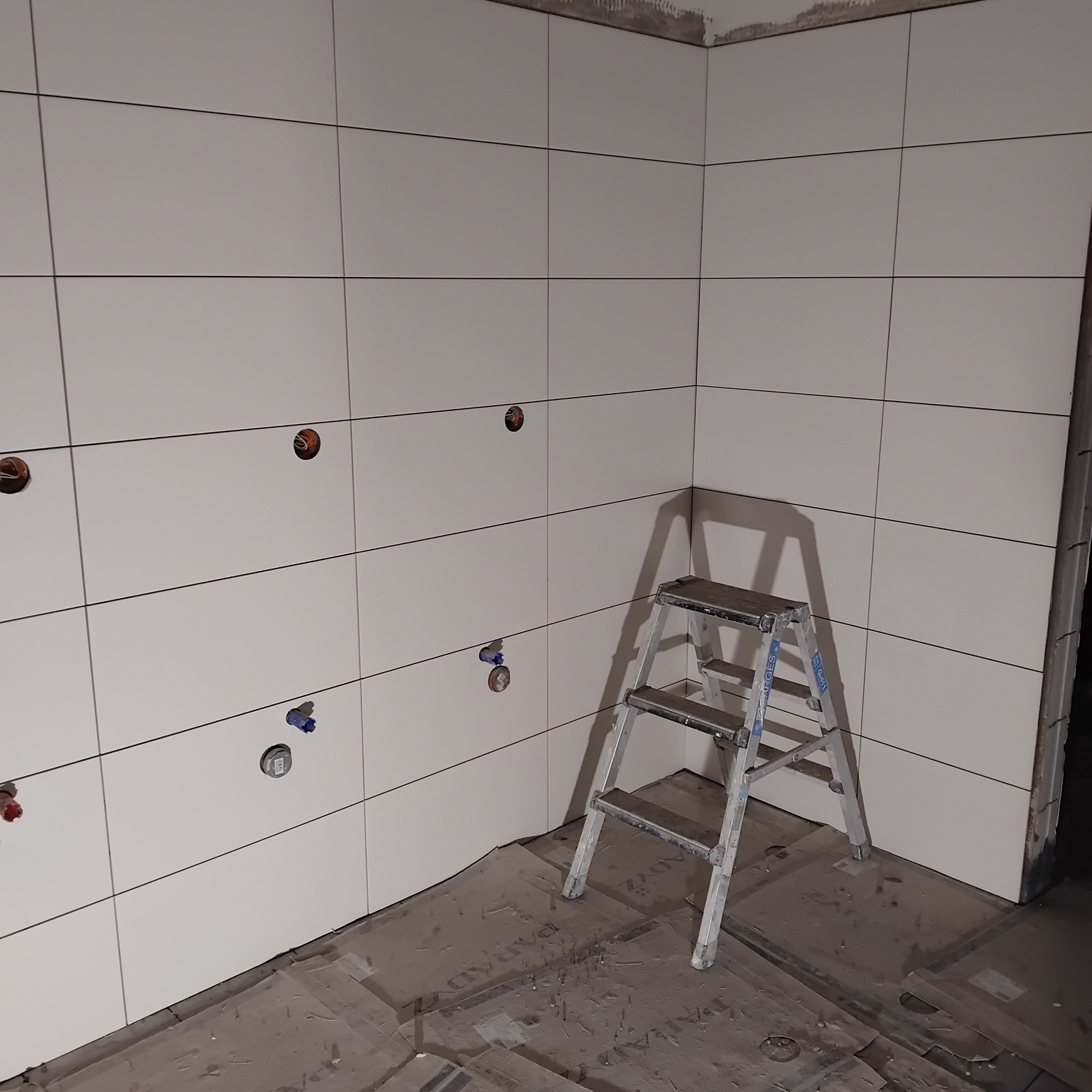 Białe, kwadratowe płytki na ścianach łazienki, z otworami na instalacje. Aluminiowa drabina stoi w rogu. Podłoga zabezpieczona kartonem.