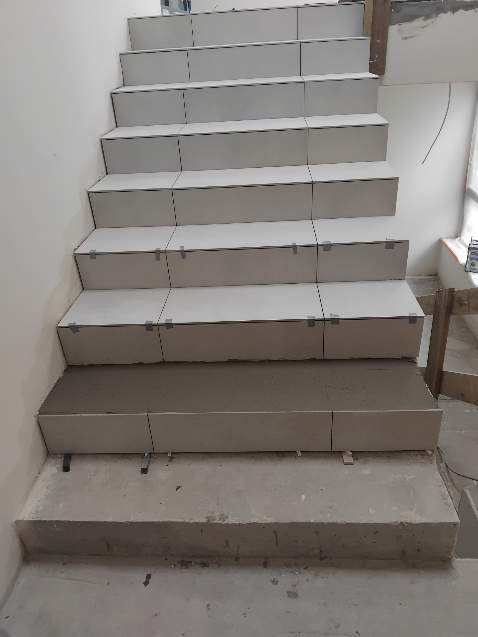 Schody w trakcie wykańczania, z jasnymi płytkami przyklejonymi do stopni, zabezpieczone taśmą. Widoczne elementy montażowe i resztki zaprawy.