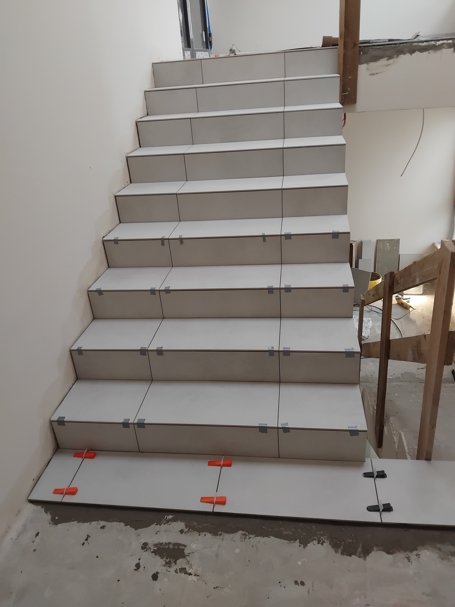 Wyłożone jasnymi płytkami schody w trakcie prac wykończeniowych. Widoczne kliny dystansowe i taśma mocująca płytki. Fragment rusztowania w tle.