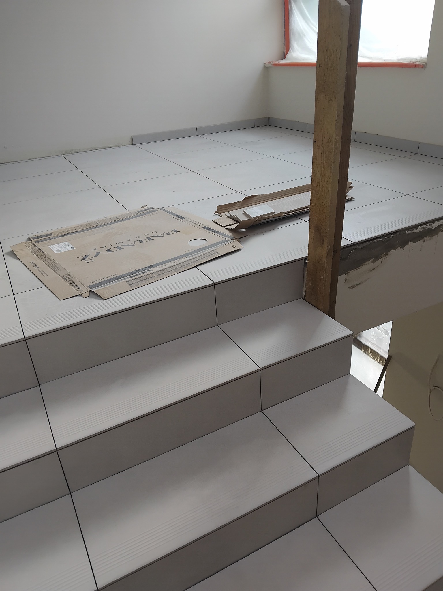 Wyłożone jasnymi płytkami schody wewnątrz budynku. Widoczna kartonowa okładzina płytek marki Paradyż. Fragment drewnianej podpory. Płytki ułożone na podłodze i schodach.