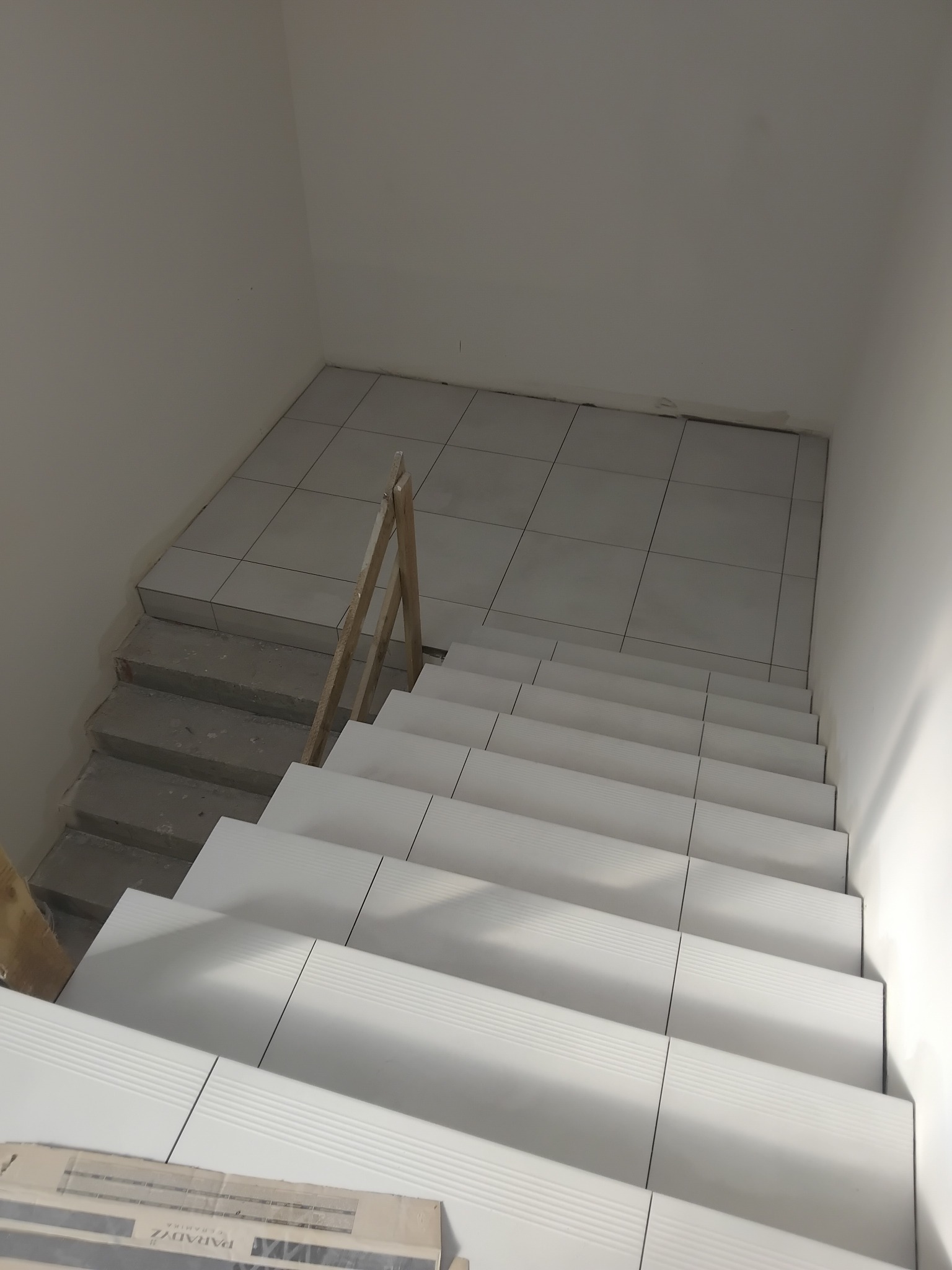 Wykończone schody z białymi płytkami z delikatnym wzorem, widok z góry. Część schodów i podest na górze wyłożone. Na dole paczka płytek.