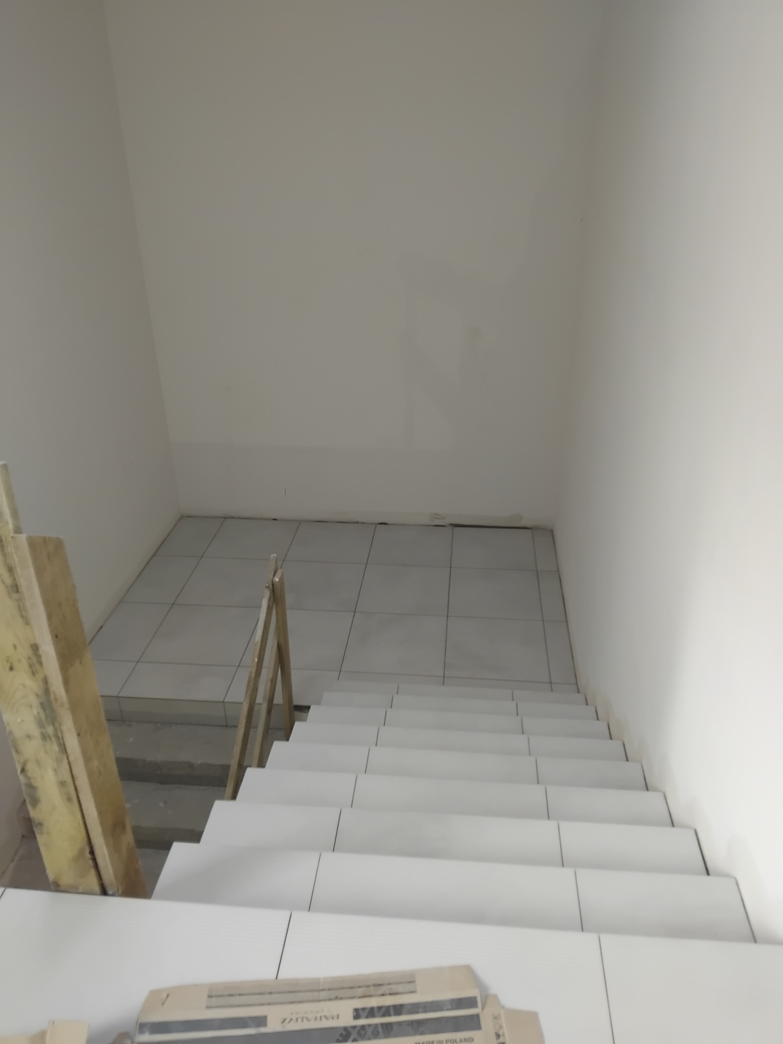 Wyłożone białymi płytkami schody w klatce schodowej. Widoczne drewniane elementy zabezpieczające. Na dole opakowanie płytek. Ujęcie z góry.