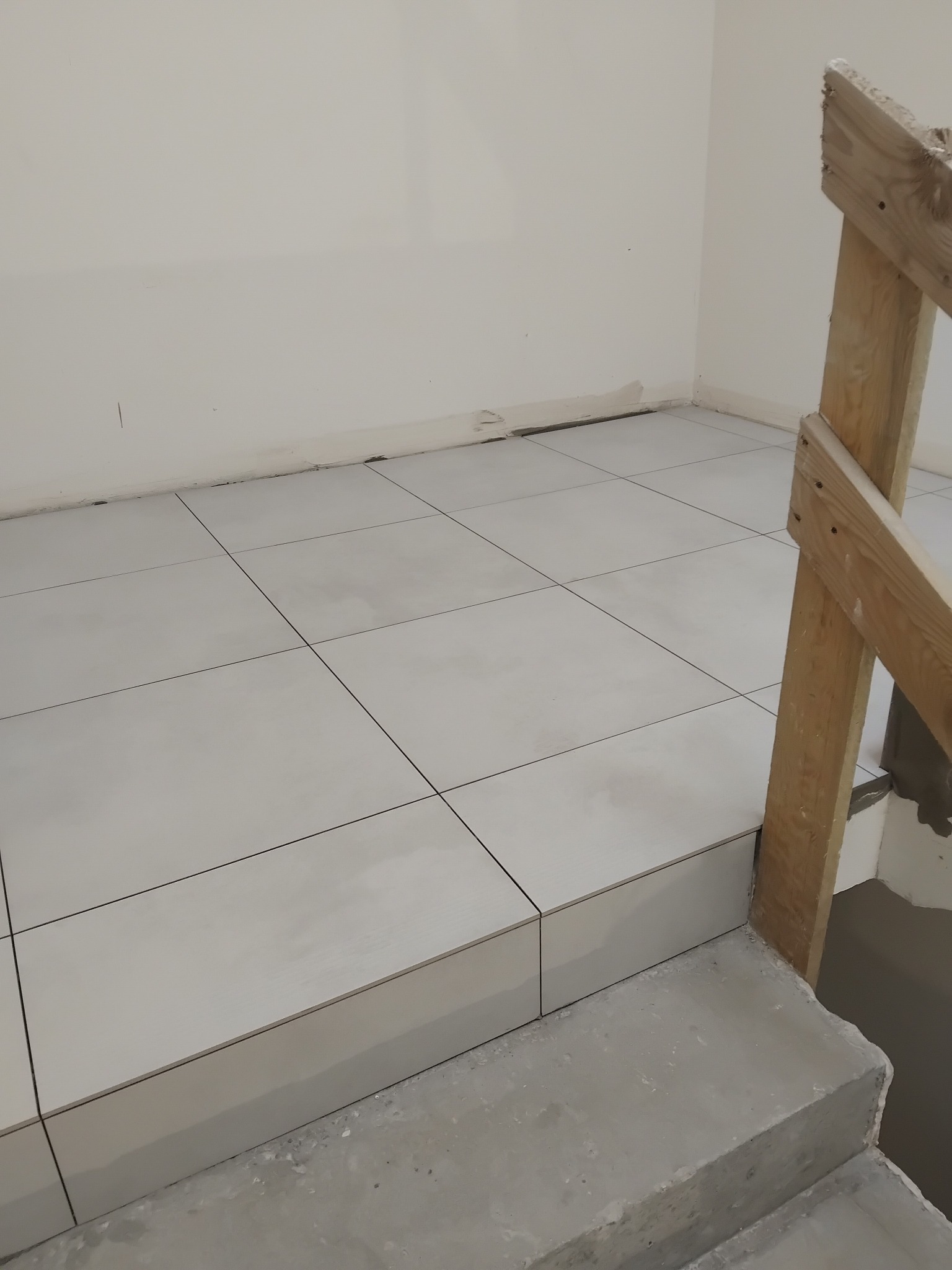 Wykończone płytki podłogowe w jasnym odcieniu szarości, ułożone na schodach i podłodze. Widoczna drewniana barierka i fragment ściany z niedoskonałościami przy podłodze.