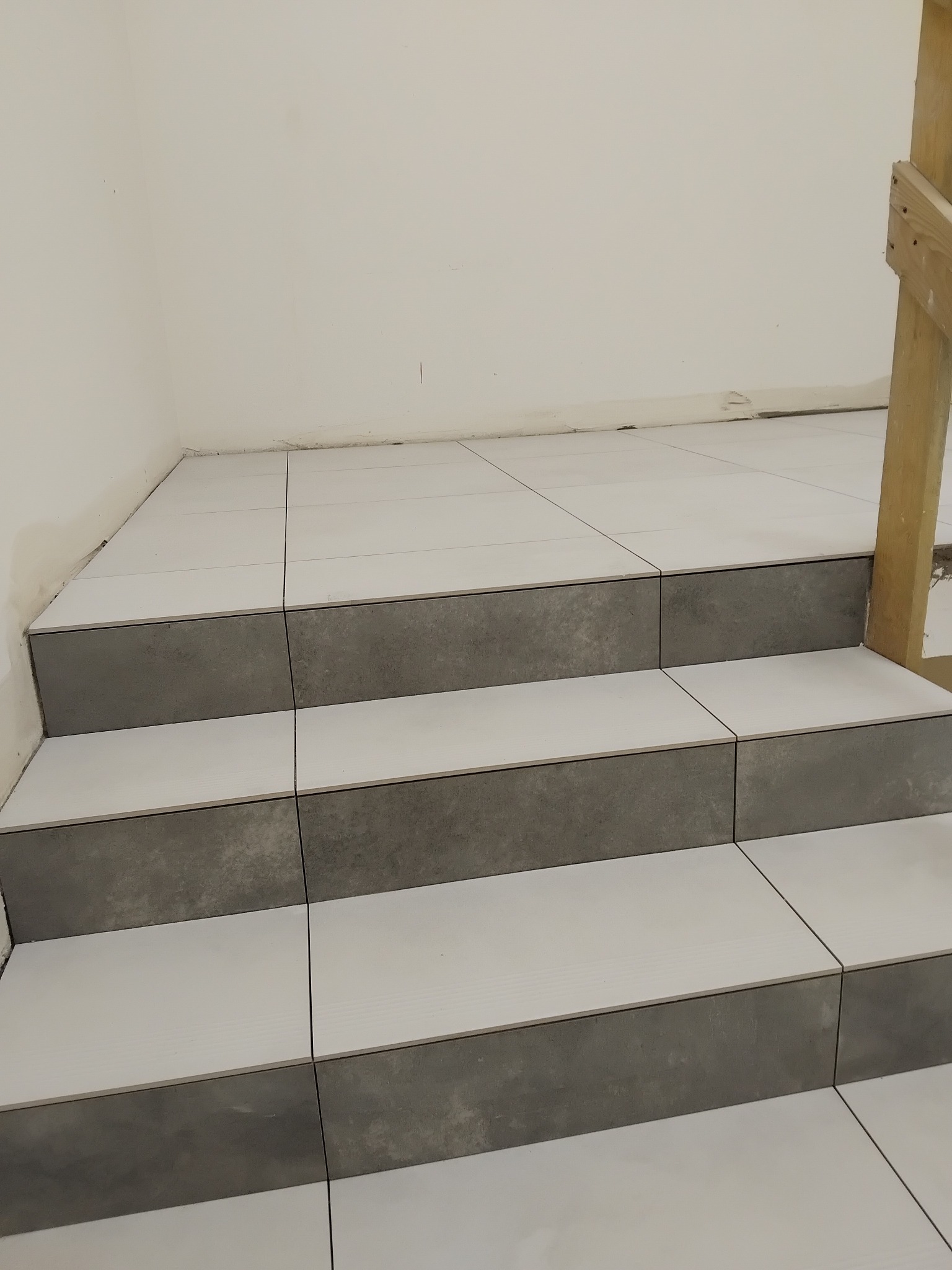 Wykończone schody z białych i szarych płytek ceramicznych, widok z dołu, fragment drewnianej konstrukcji w prawym górnym rogu. Estetyczne połączenie kolorów i materiałów.