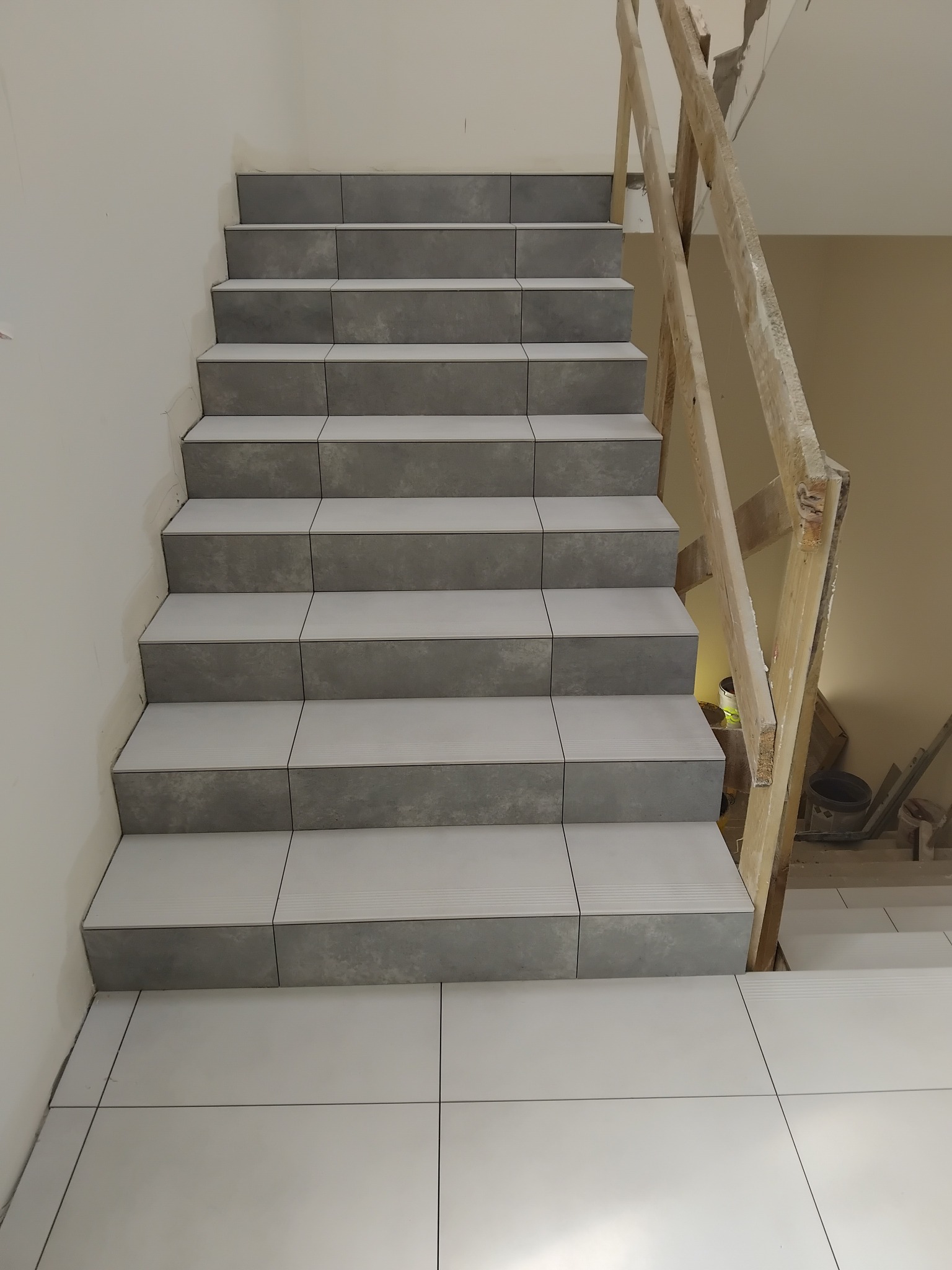 Schody wyłożone szarymi płytkami z białymi fugami. Widoczne prowizoryczne drewniane zabezpieczenie schodów. Podłoga również wyłożona płytkami.