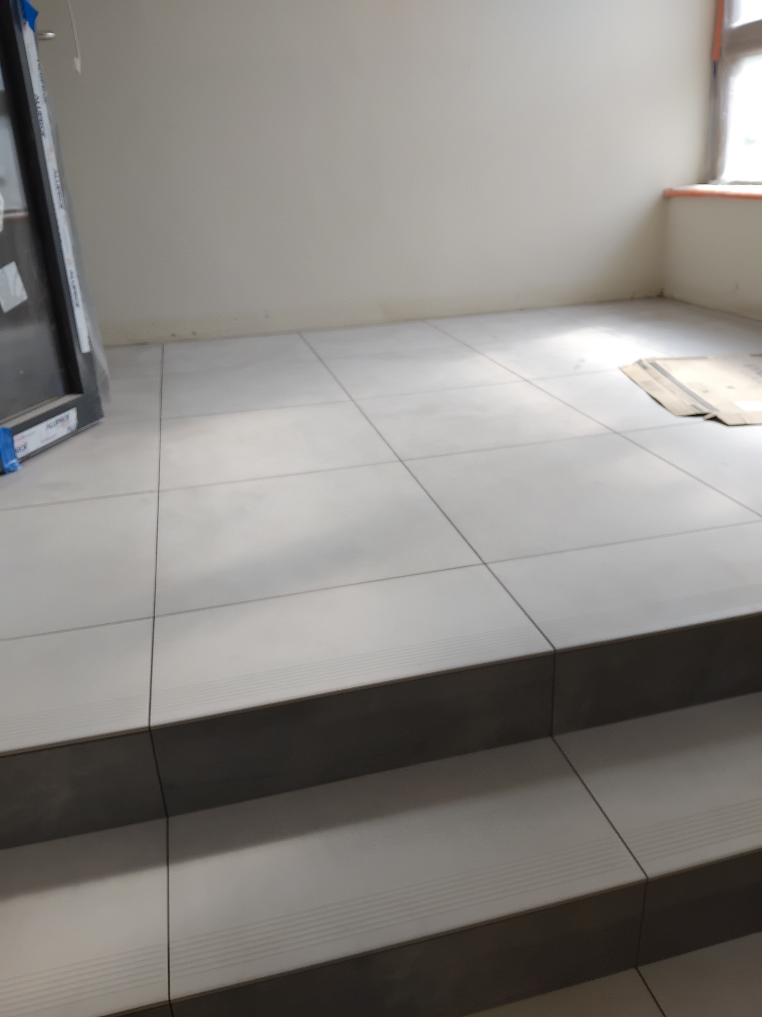 Wykończone płytki podłogowe w jasnych odcieniach szarości, układane schodkowo. Widoczne fugi i fragment okna z parapetem. Częściowo widoczne drzwi.