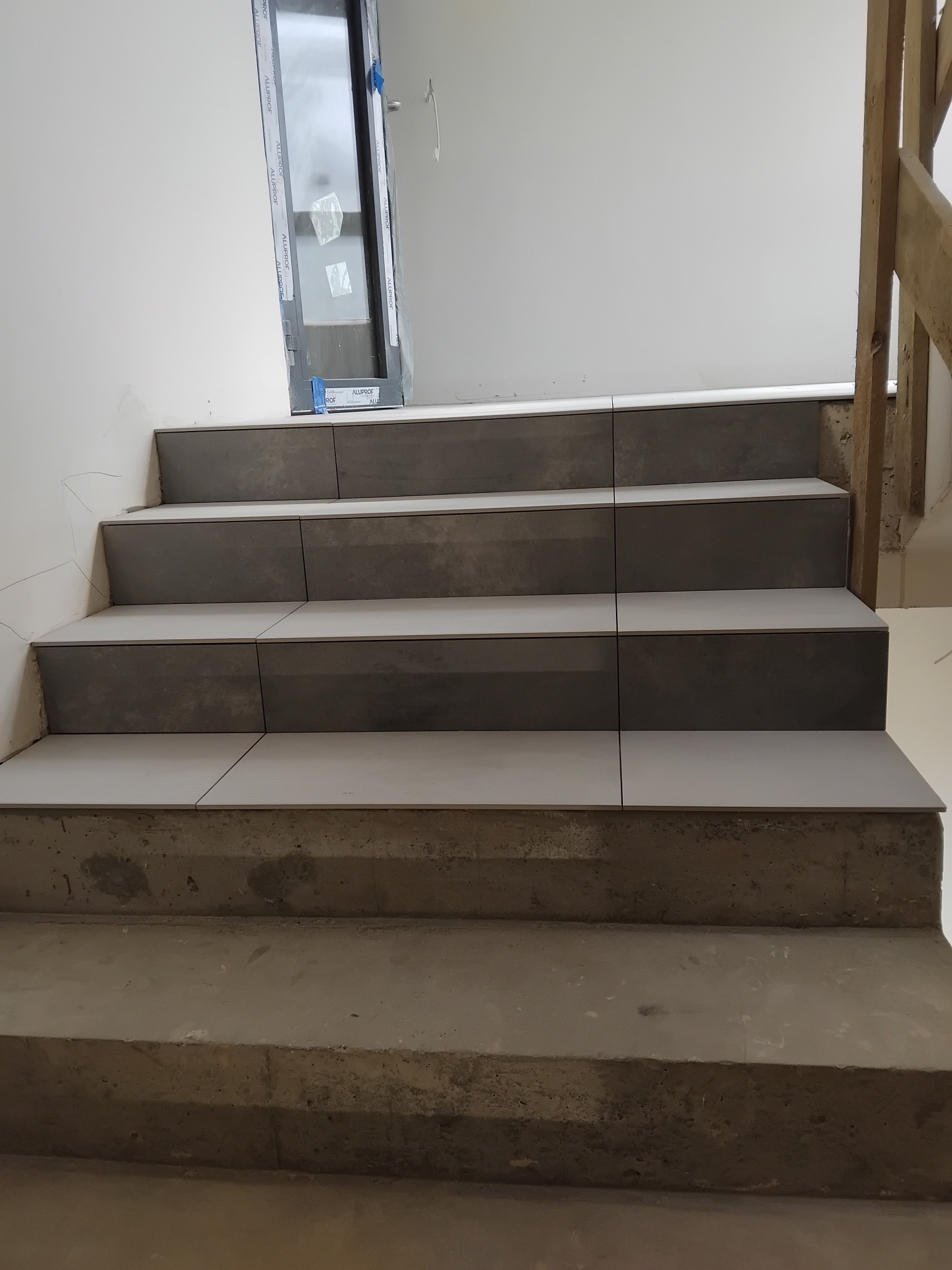 Wykończone płytkami schody w szarościach, widoczne niedokończone ściany i okno. Układ płytek prosty, minimalistyczny. Etap prac wykończeniowych.