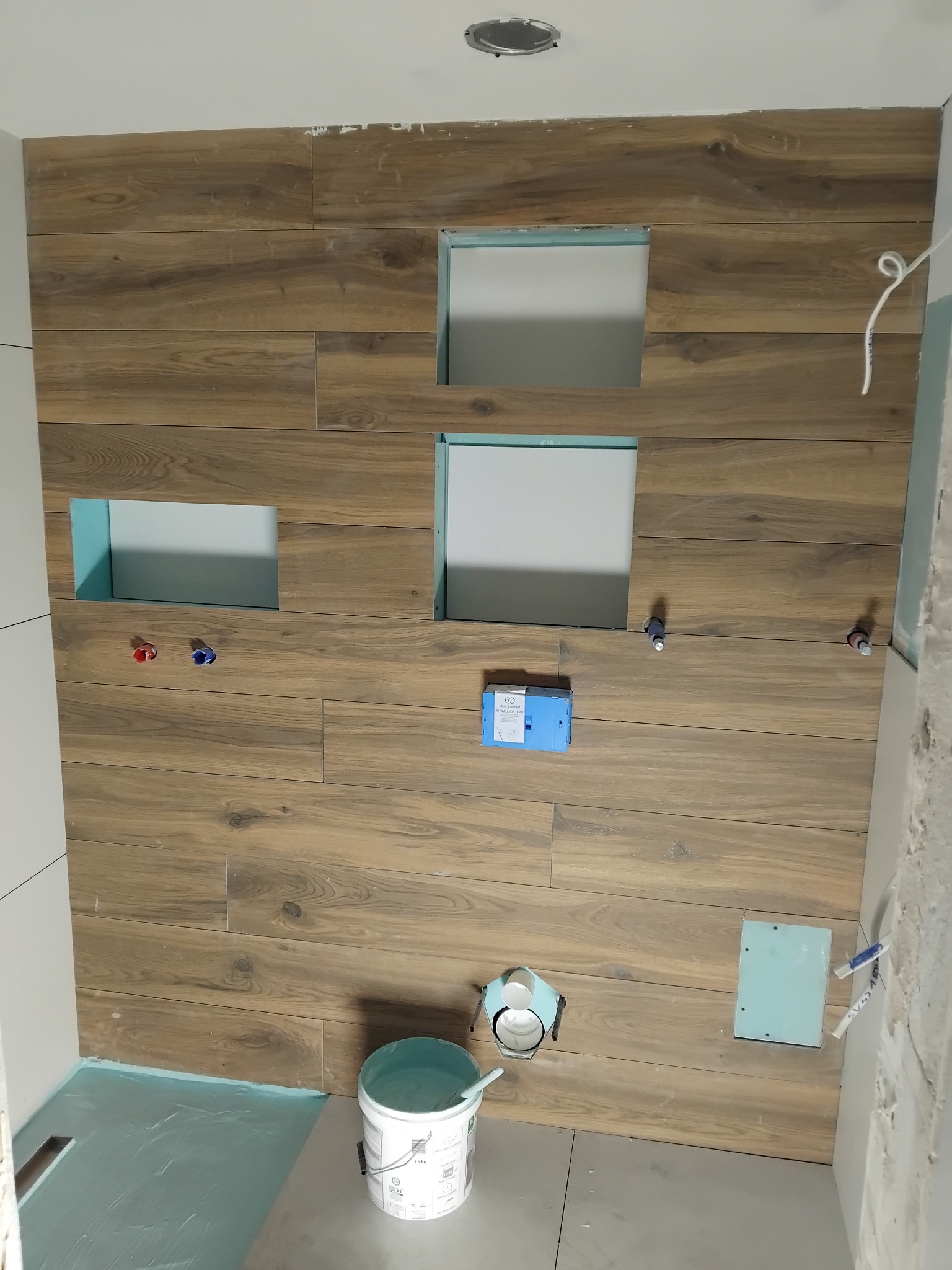 Ściana w łazience wyłożona płytkami imitującymi drewno z otworami na instalacje i wnękami, widoczne narzędzia i wiadro z klejem.