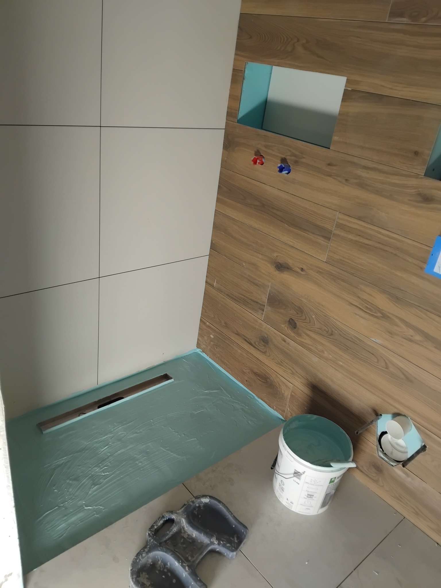 Fragment łazienki w trakcie prac: białe płytki ścienne, drewnopodobne płytki na ścianie z wnękami, brodzik z odpływem liniowym, wiadro z zaprawą i kielnia.