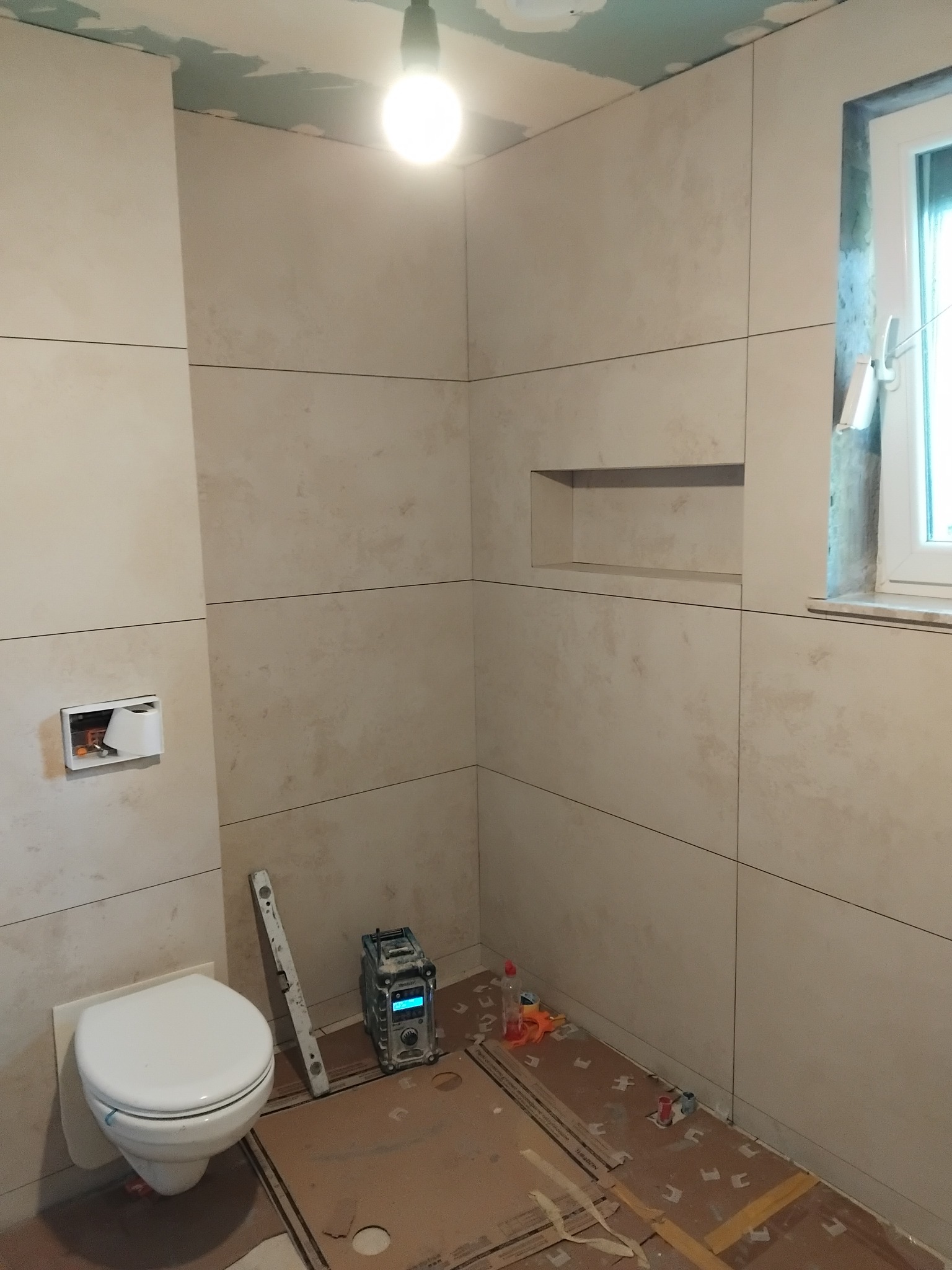 Wykończona beżowa łazienka z podtynkową toaletą i wnęką w ścianie. Na podłodze karton zabezpieczający, obok poziomica i laser krzyżowy.