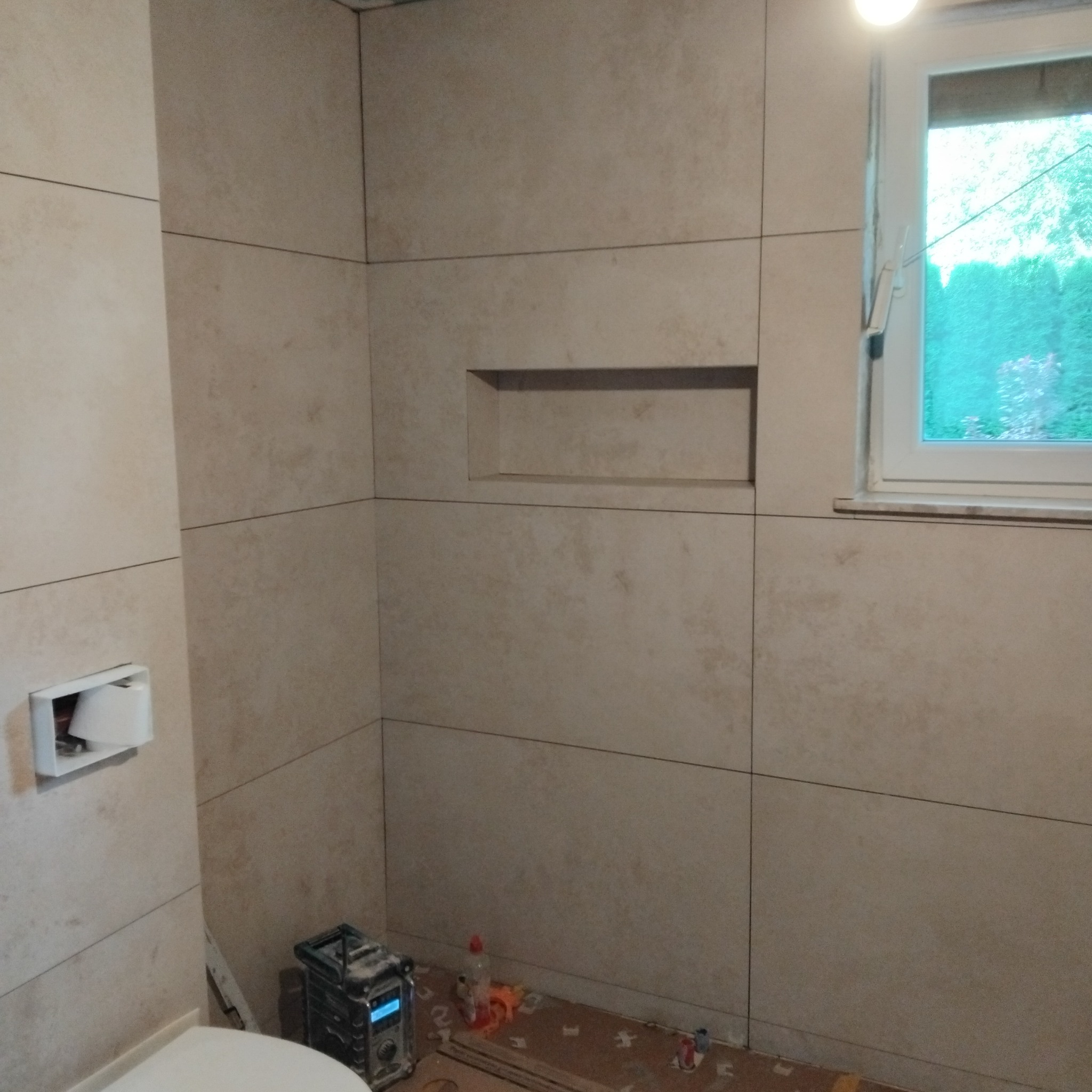 Wykończona ściana w łazience z beżowych płytek, z wnęką i oknem. Na podłodze widoczne ślady po pracach glazurniczych i narzędzia.