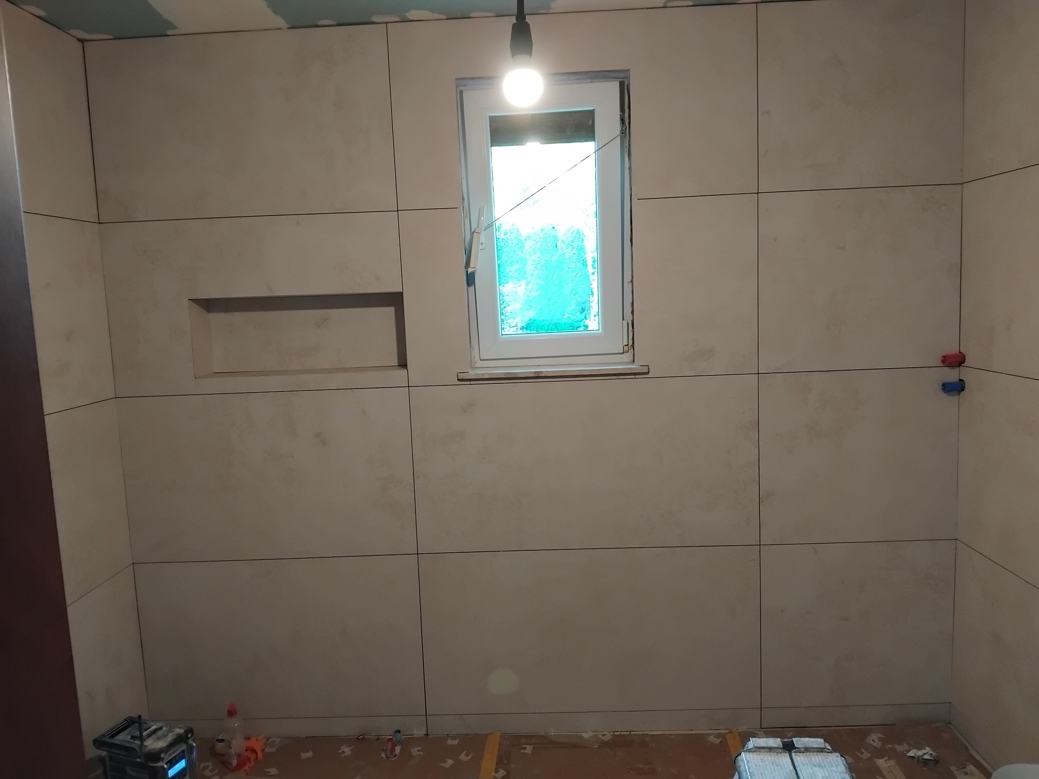 Wyłożona beżowymi płytkami łazienka z oknem. Widoczne narzędzia glazurnicze na podłodze i wnęka w ścianie. Górna część ściany nie wykończona.