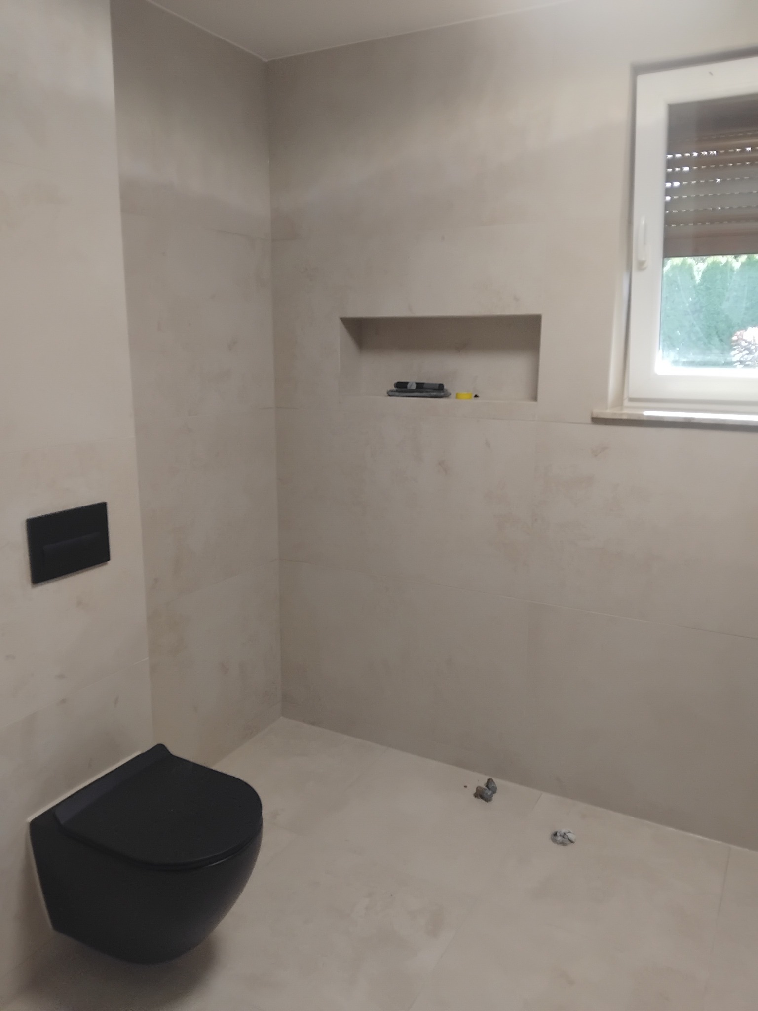 Minimalistyczna łazienka w Połczyn-Zdrój z beżowymi płytkami, czarną toaletą podwieszaną i wnęką. Widoczne narzędzia glazurnicze.