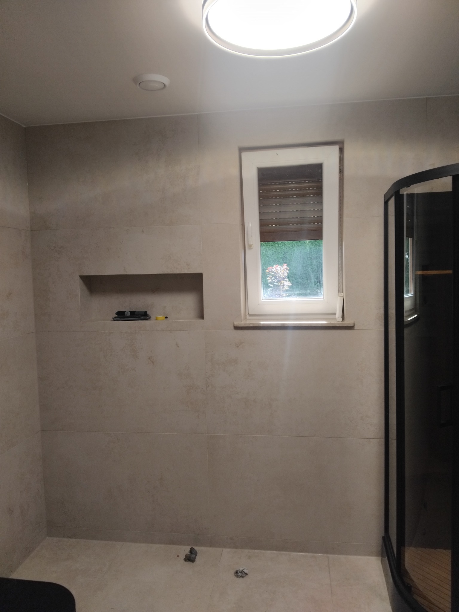 Beżowa łazienka z oknem i kabiną prysznicową. Na ścianie wnęka z narzędziami. Widoczny fragment kabiny prysznicowej z czarnymi profilami.