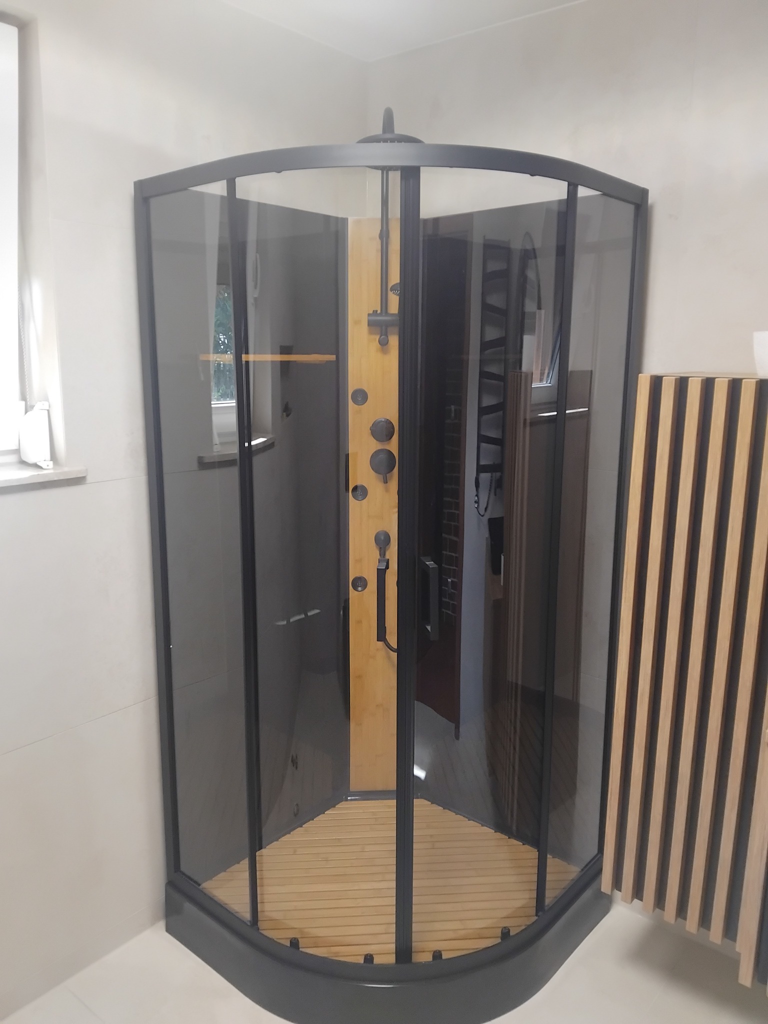 Kabina prysznicowa z ciemnymi ramami i drewnianym panelem w środku, widoczna w łazience z jasnymi płytkami i elementami dekoracyjnymi z drewna.