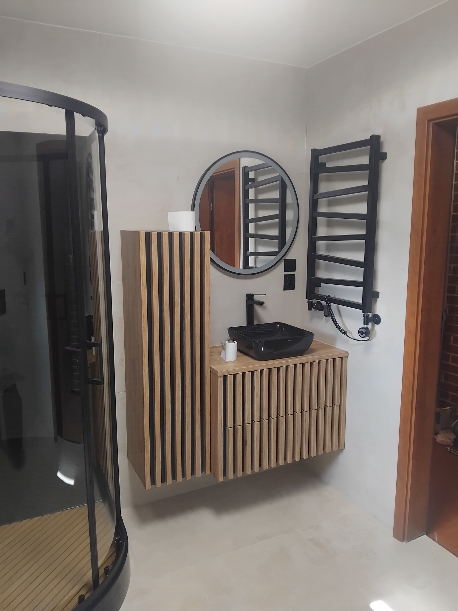 Nowoczesna łazienka z czarną armaturą i drewnianymi lamelami na szafce umywalkowej oraz grzejniku. Kabina prysznicowa z czarnymi profilami.