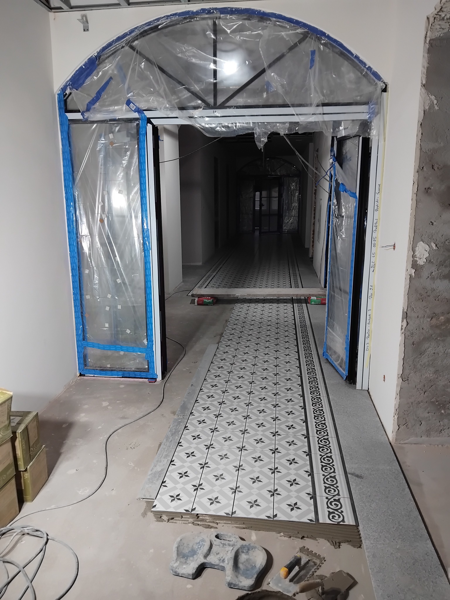 Kładzenie wzorzystych płytek podłogowych w korytarzu, zabezpieczone folią drzwi łukowe, narzędzia glazurnicze w dolnym rogu. Etap prac wykończeniowych.