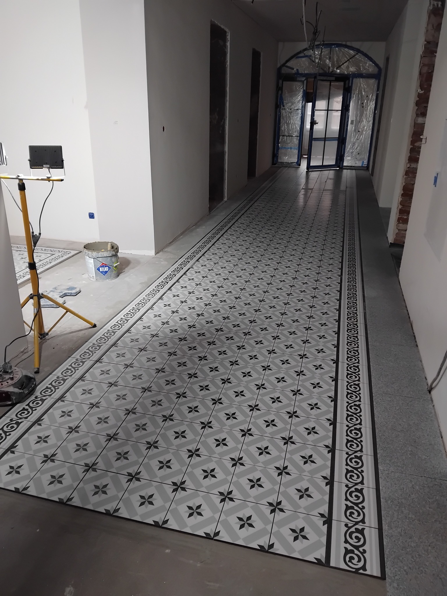 Ułożona, wzorzysta, szaro-biała podłoga w korytarzu, z widocznym wiadrem z klejem i lampą budowlaną. W tle przeszklone drzwi i surowy stan wykończenia ścian.