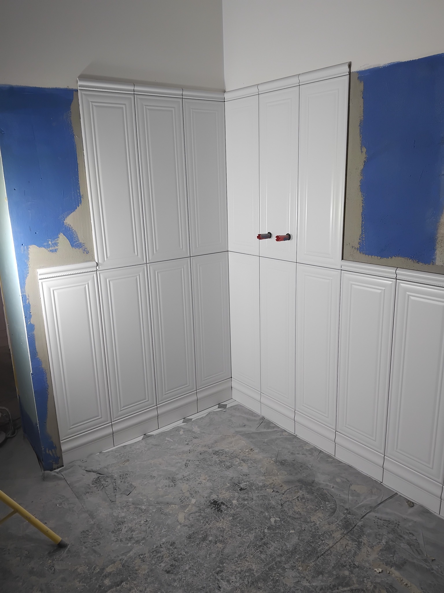 Białe, kasetonowe płytki ścienne w trakcie montażu w narożnej łazience. Widoczne nieotynkowane fragmenty ścian i surowa podłoga. Dwa wystające zawory.