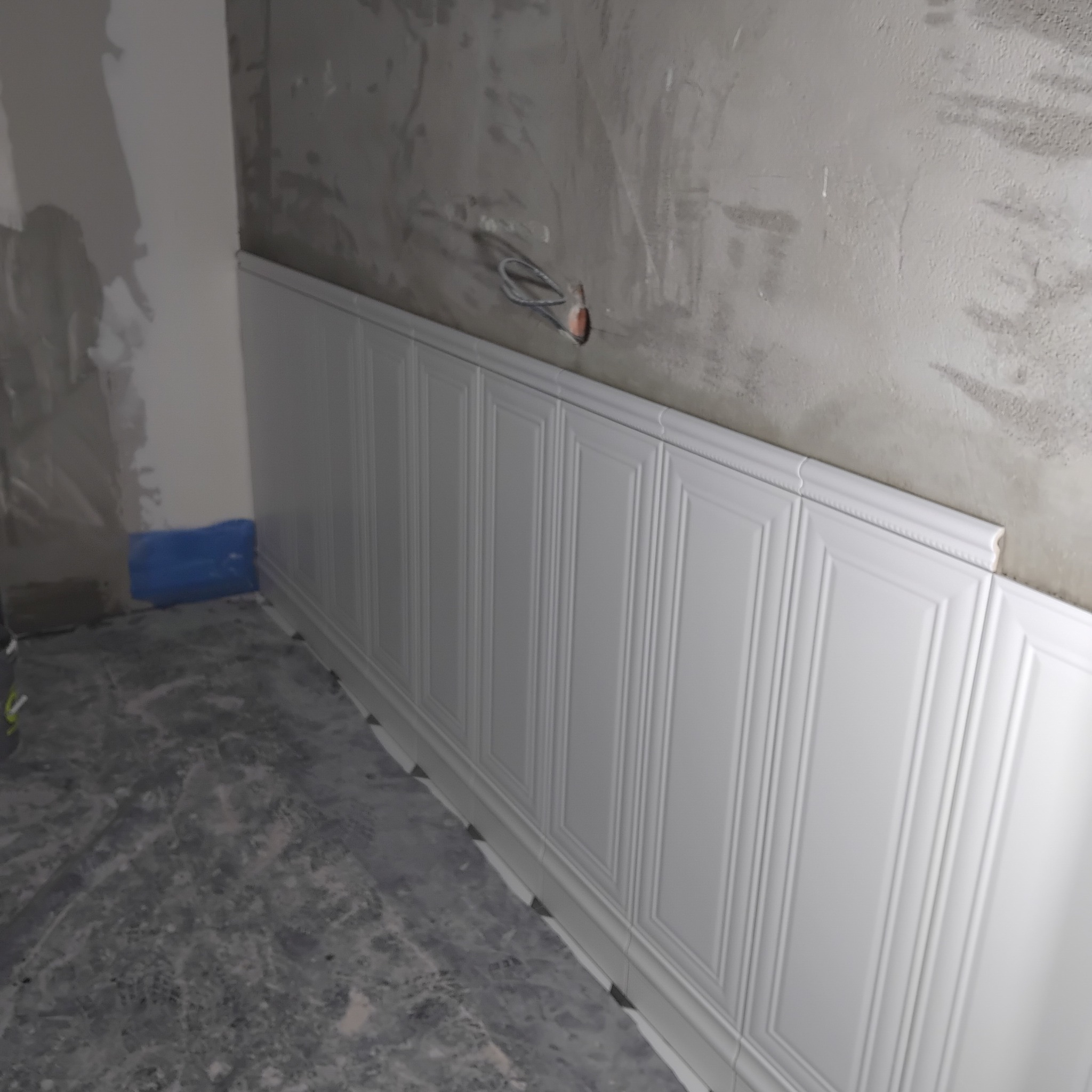 Montaż białej boazerii angielskiej na ścianie w surowym wnętrzu. Widoczne przewody elektryczne i zabezpieczenie podłogi folią. Prace wykończeniowe w toku.