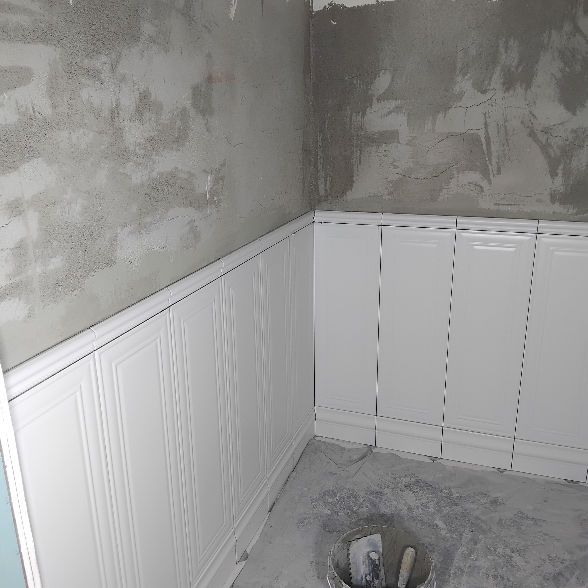 Białe, panelowe płytki ścienne w trakcie montażu, widoczne surowe ściany i wiadro z narzędziami, ukazujące etap prac wykończeniowych w pomieszczeniu.