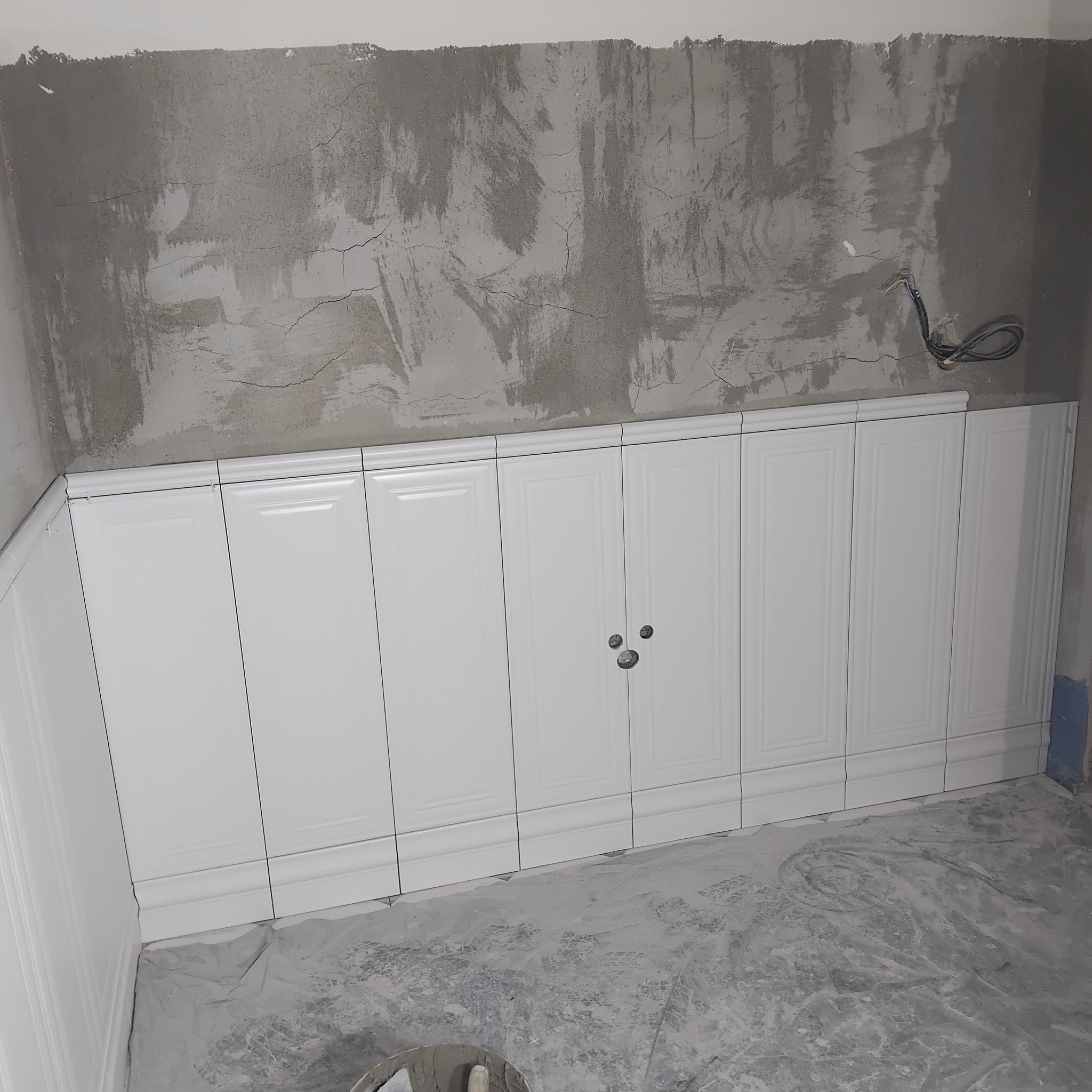 Białe, panelowe płytki ścienne w trakcie montażu, widoczne przewody elektryczne i surowy beton na ścianie powyżej. Podłoga pokryta zaprawą.