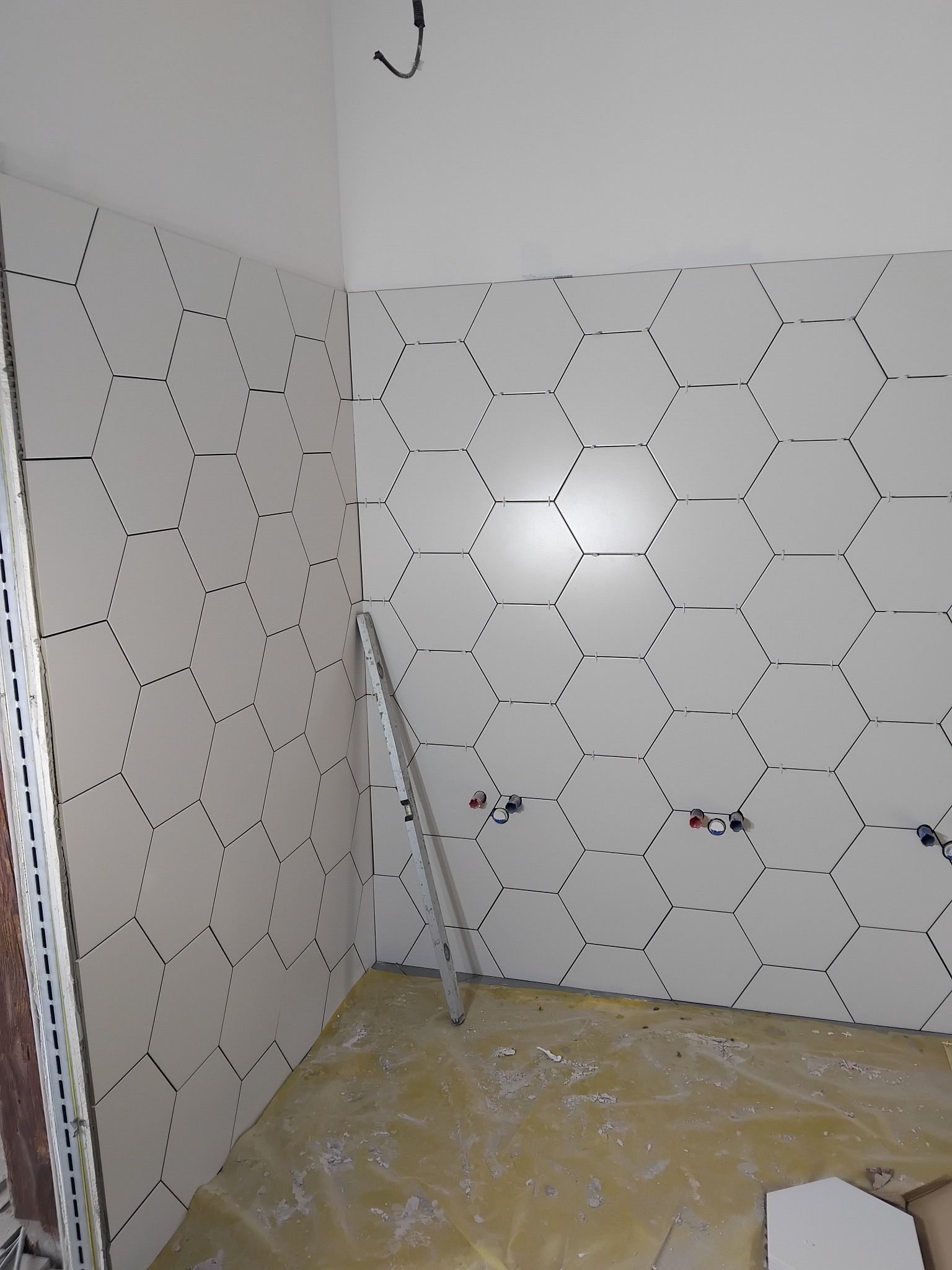 Układanie heksagonalnych, białych płytek na ścianie łazienki, widoczne otwory na instalacje, poziomica oparta o ścianę. Prace glazurnicze w toku.