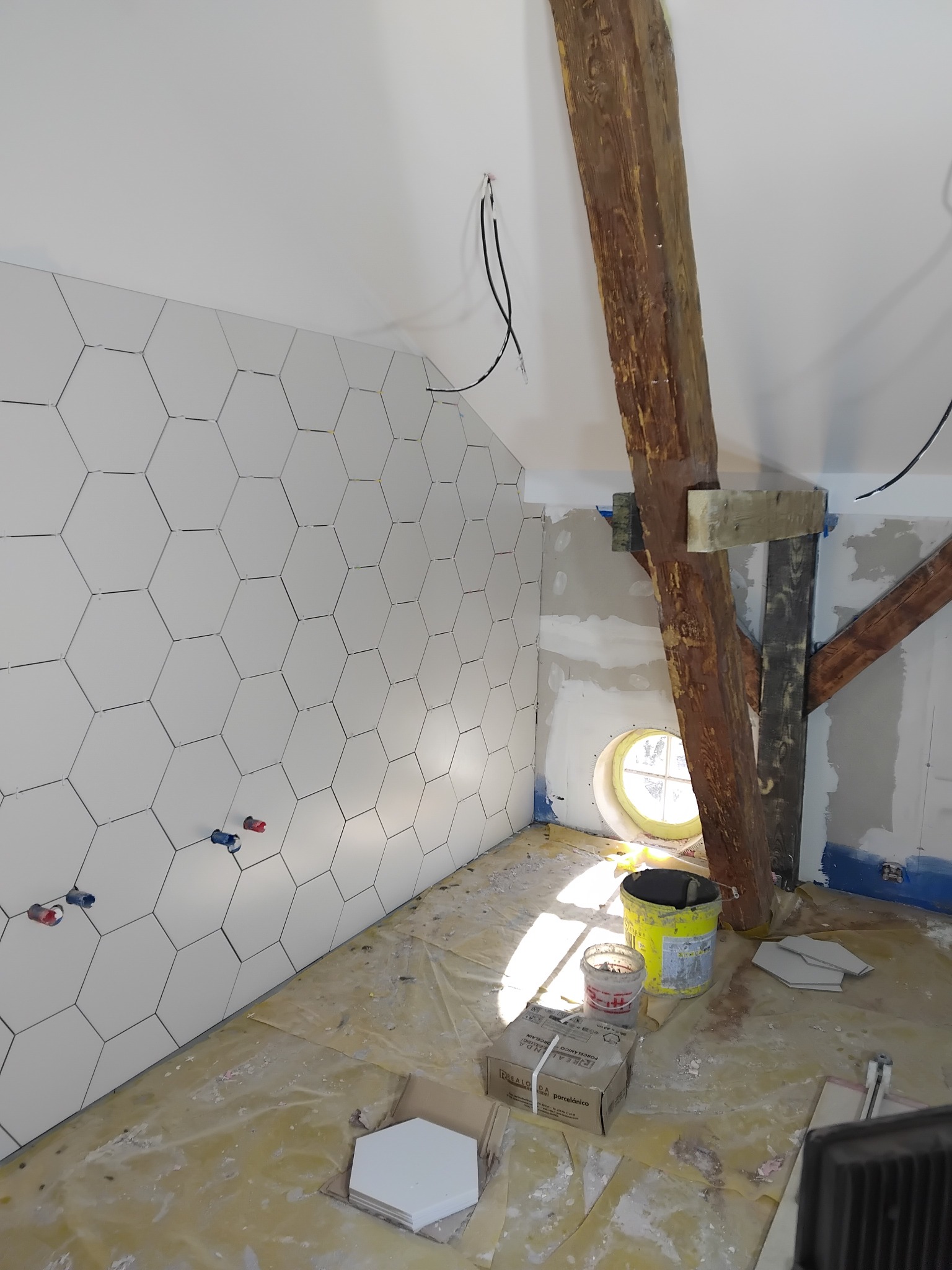 Ściana w łazience z heksagonalnymi, białymi płytkami, widoczne wystające rury, drewniana belka i okrągłe okno. Podłoga zabezpieczona folią, widoczne narzędzia i materiały.