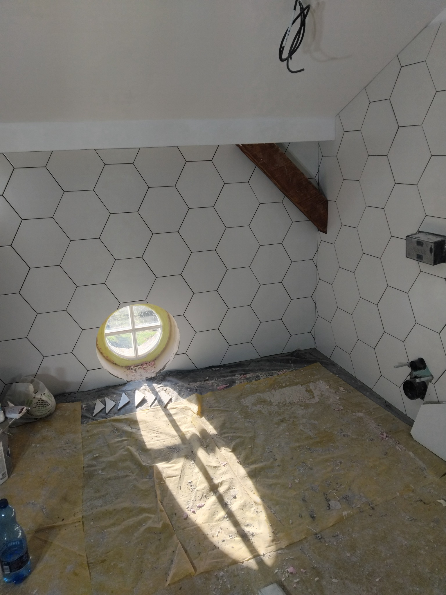 Łazienka w trakcie wykańczania: heksagonalne płytki na ścianach, okrągłe okno, zabezpieczona podłoga. Widoczne elementy instalacji i narzędzia.