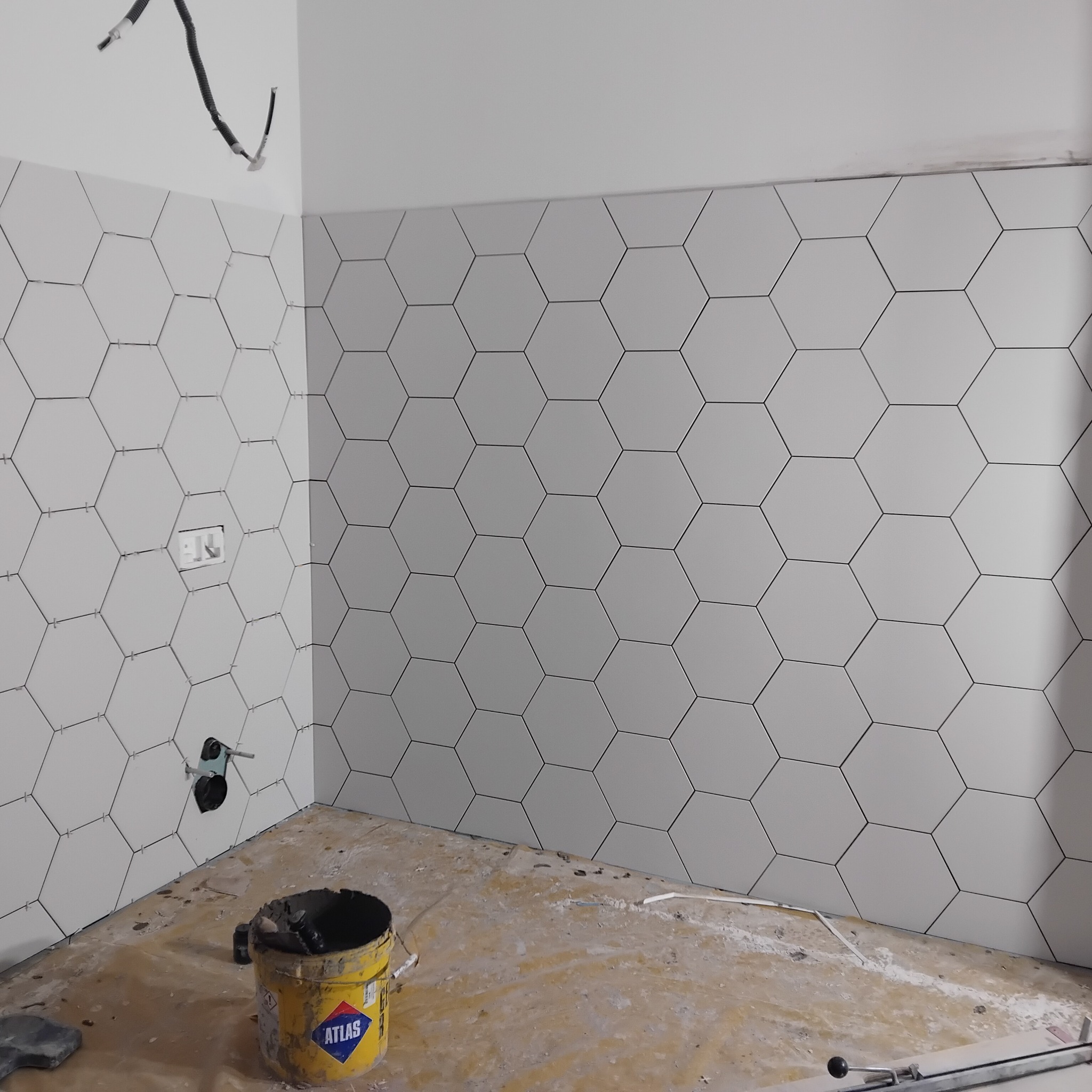Ściana wyłożona białymi, heksagonalnymi płytkami z ciemną fugą, widoczny fragment instalacji i wiadro z klejem Atlas, podłoga przygotowana pod dalsze prace.