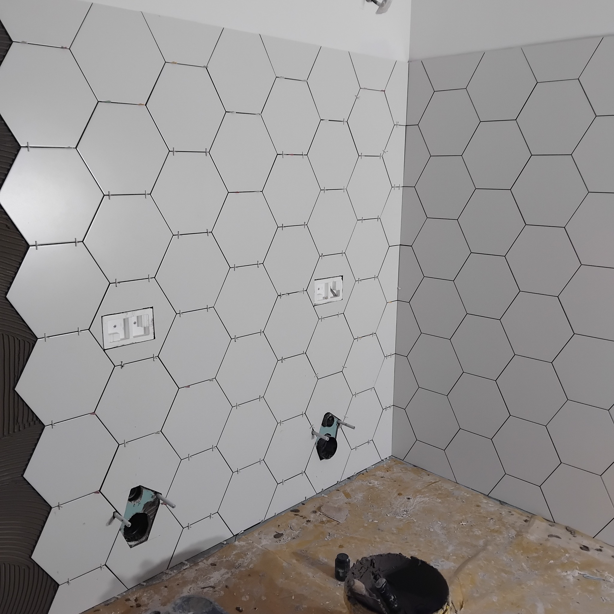 Ściana z białych, heksagonalnych płytek, częściowo ułożonych, z widocznymi otworami na instalacje i krzyżykami dystansowymi. W tle wiadro z zaprawą.