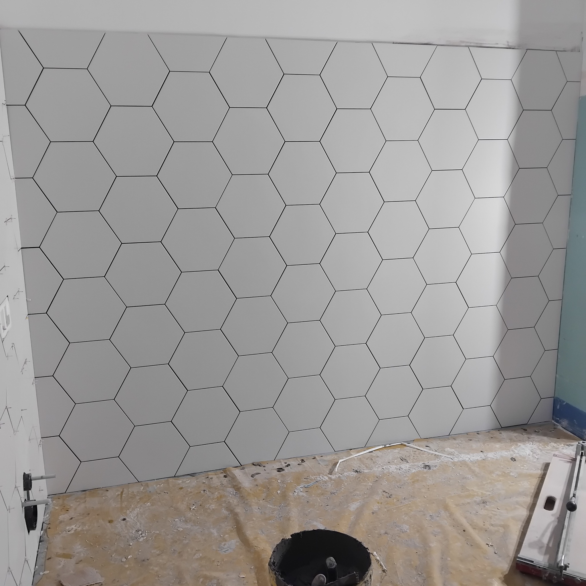 Ściana wyłożona białymi, heksagonalnymi płytkami w łazience w trakcie remontu. Widoczny fragment podłogi z zabrudzeniami i narzędzia glazurnicze.
