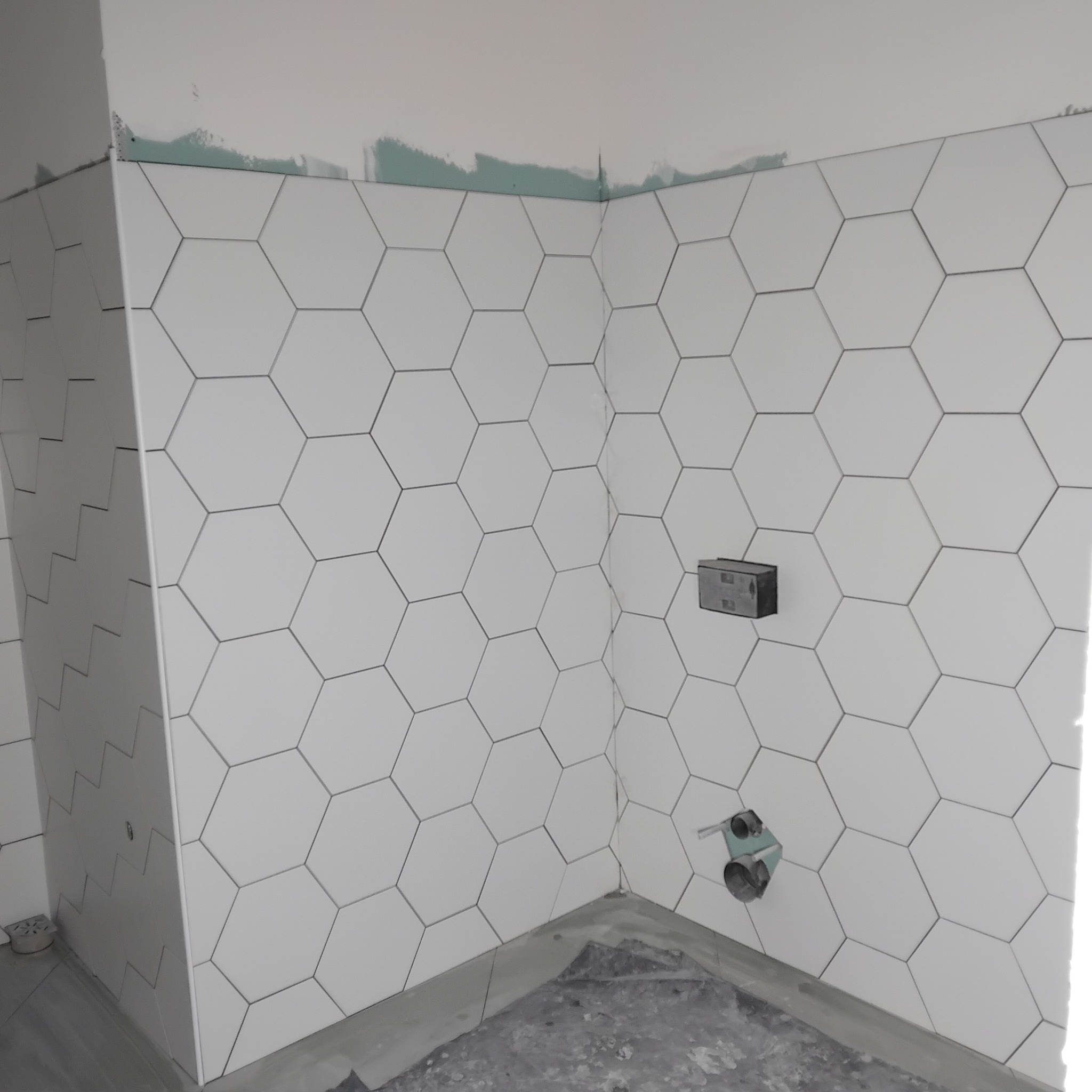 Ściana łazienki wyłożona białymi, heksagonalnymi płytkami. Widoczne wystające elementy instalacji wodnej i elektrycznej. Częściowo wykończone górne krawędzie ścian.