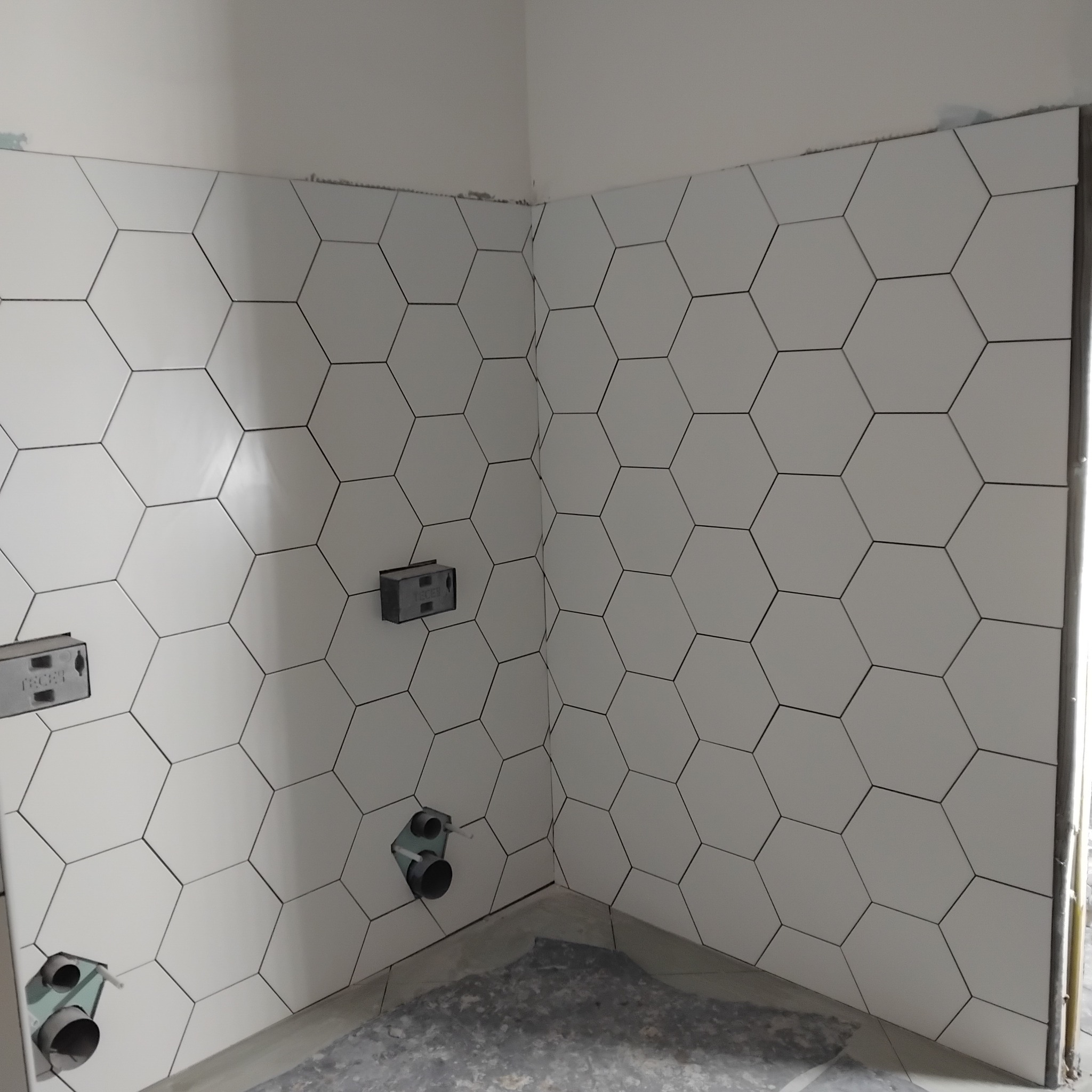 Ściana wyłożona białymi, heksagonalnymi płytkami z czarną fugą, widoczne przyłącza wodne i elektryczne. Ukończony etap prac w łazience.