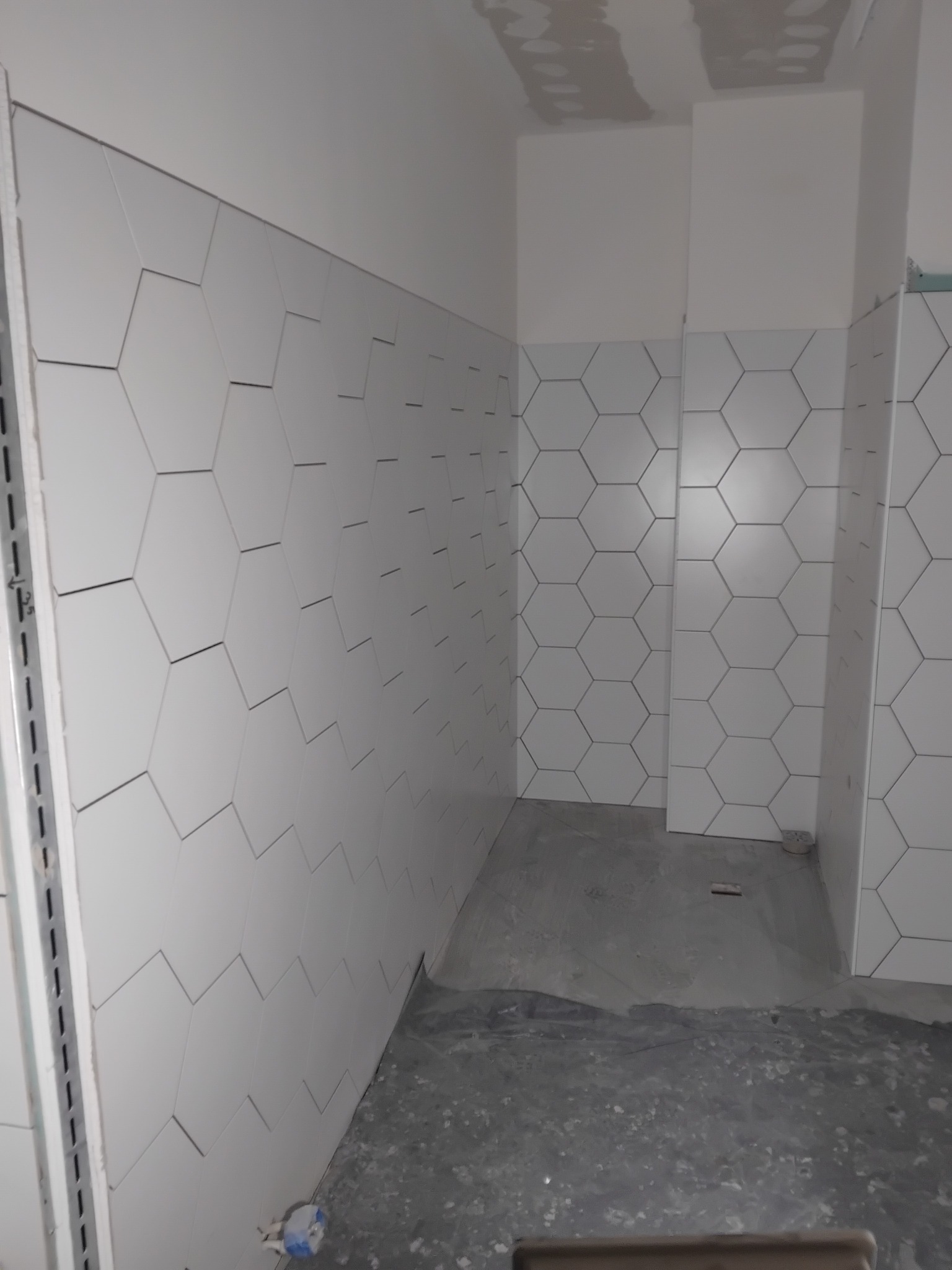 Ściana wyłożona białymi, heksagonalnymi płytkami w łazience, z widoczną szarą podłogą i fragmentem sufitu. Płytki ułożone w regularny wzór, z ciemnymi fugami.