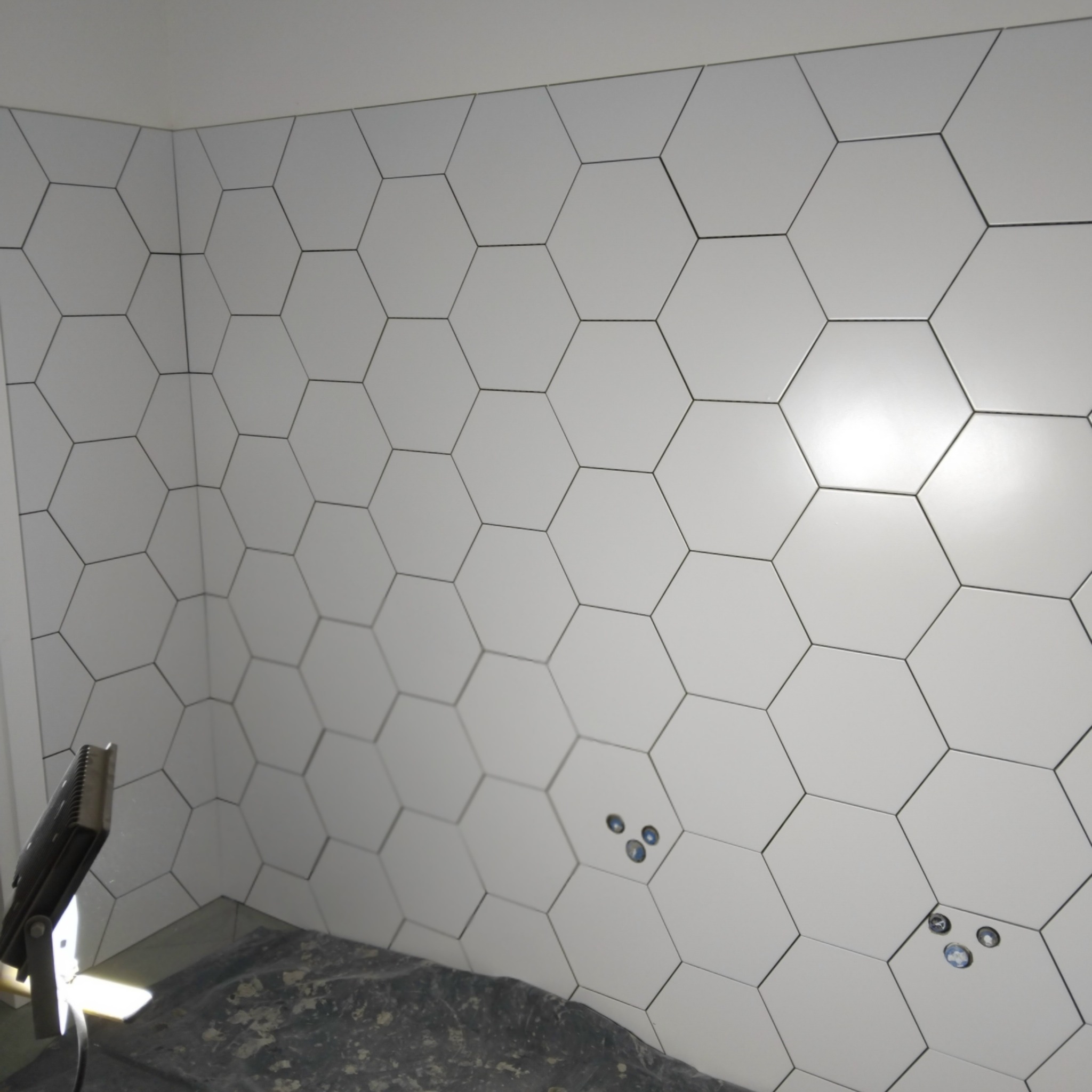 Ściana wyłożona białymi, heksagonalnymi płytkami, widoczne otwory na instalacje. W rogu lampa budowlana. Nietypowy kształt płytek.