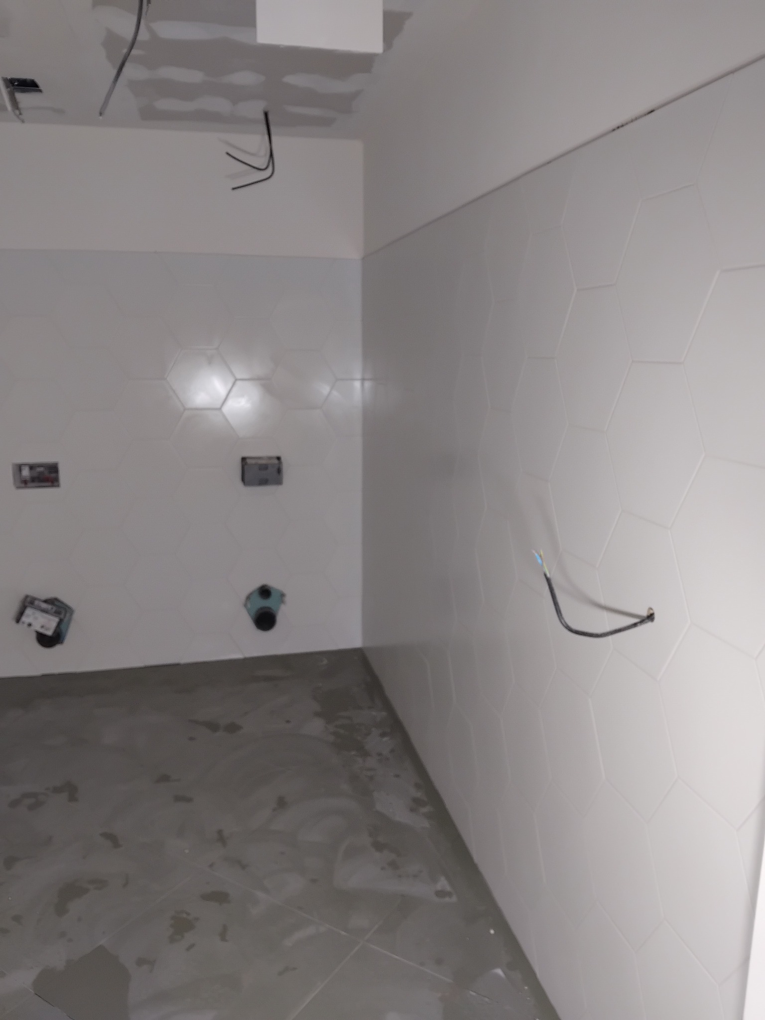 Łazienka w trakcie wykończenia: białe heksagonalne płytki na ścianach, widoczne instalacje, surowa podłoga. Etap prac przed montażem armatury i wykończeniem.