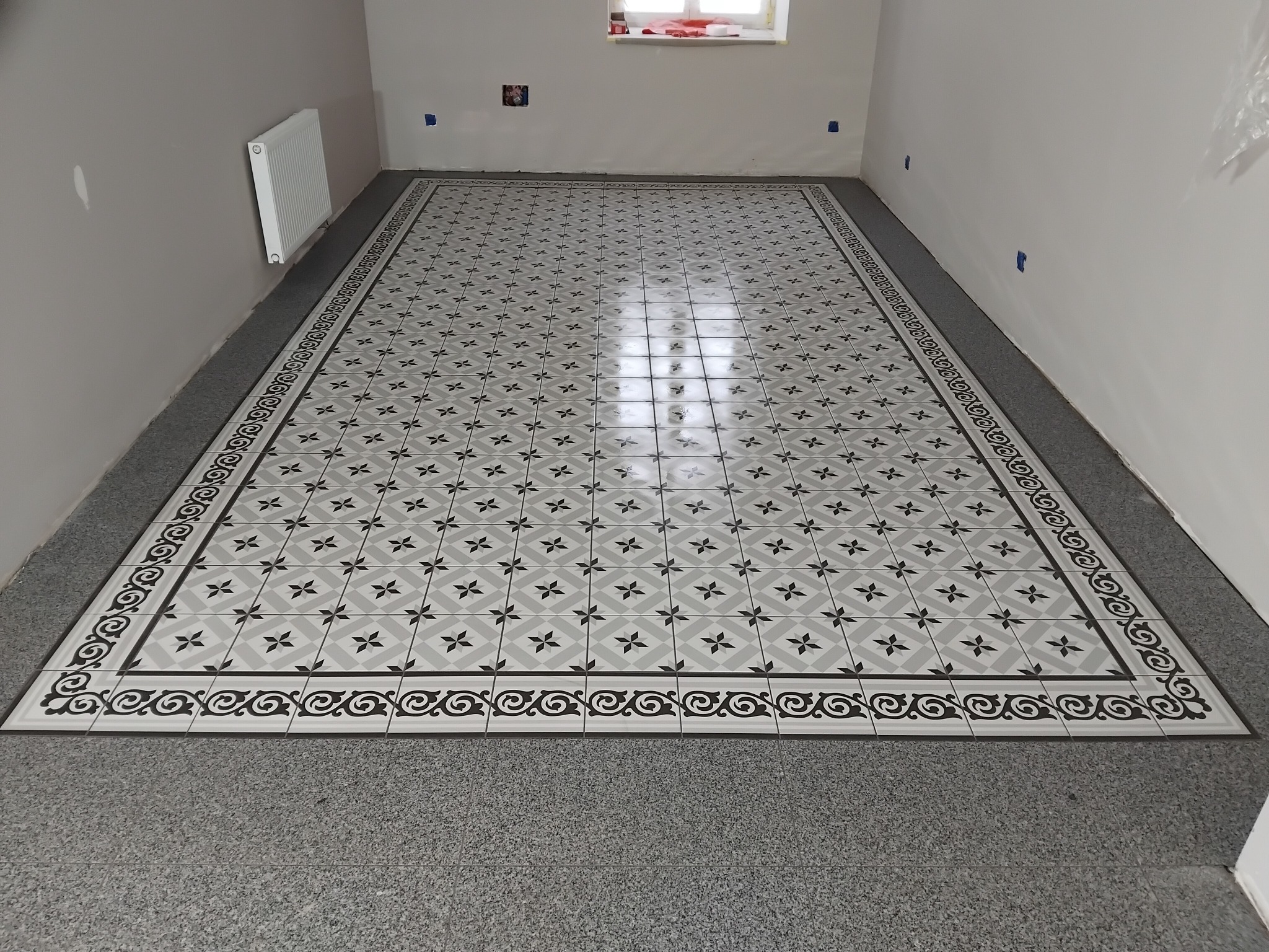 Ukończona podłoga z wzorzystych, szaro-białych płytek w nowym pomieszczeniu. Widoczne szare podłoże i białe ściany z niebieskimi oznaczeniami.