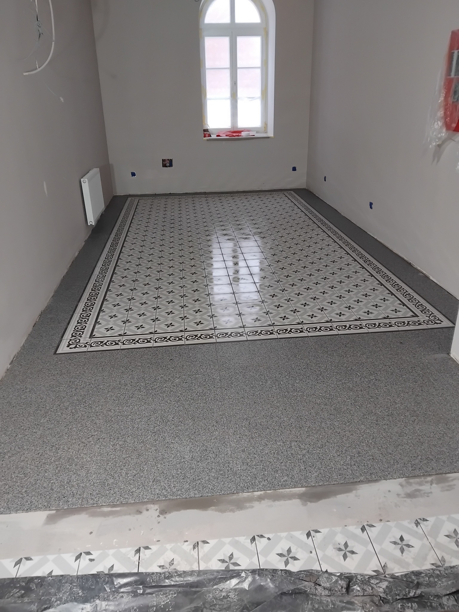 Ukończona podłoga w łazience z szarych płytek z wzorem, otoczona szarą, drobną mozaiką. Widoczne okno i grzejnik. Praca glazurnika.